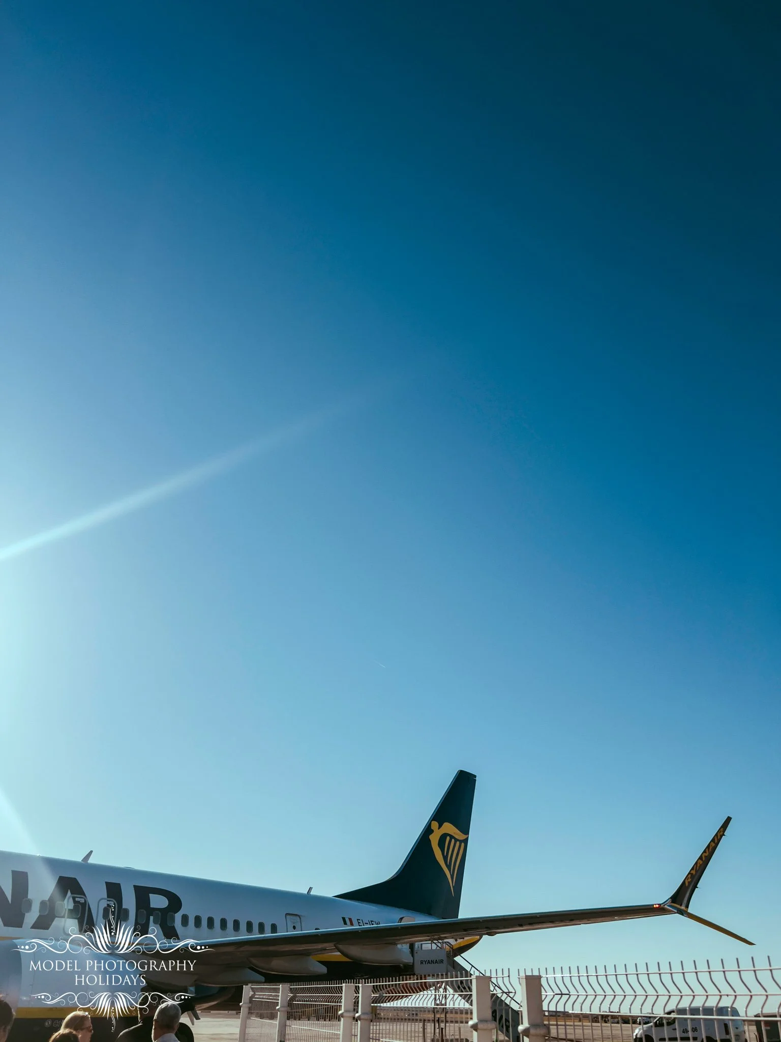 View of a Ryanair airplane at an airport with parts of the aircraft visible, including the tail with the Ryanair logo, against a clear blue sky. A fence and airport personnel are seen in the foreground.