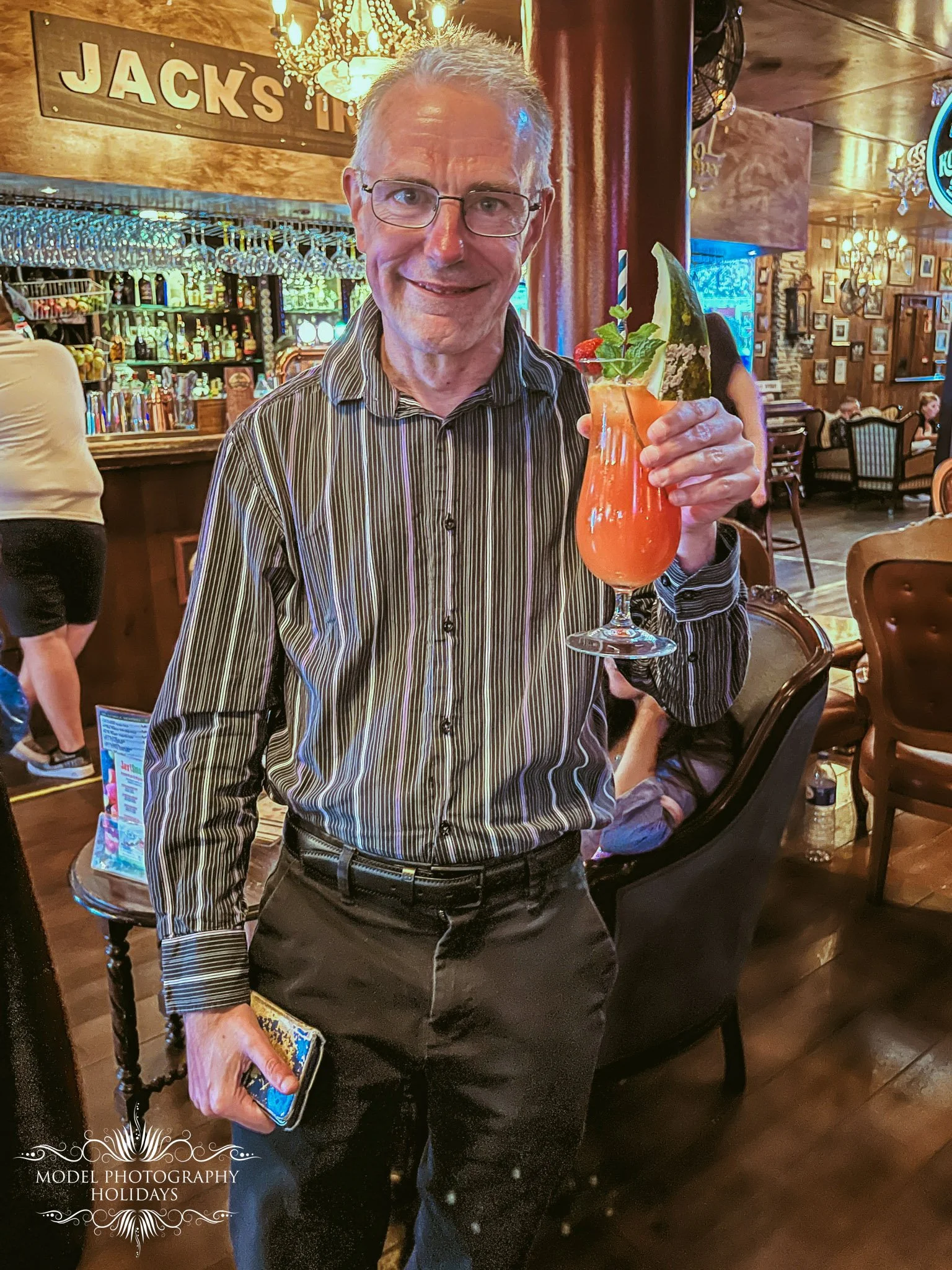 A smiling older man with glasses wearing a striped shirt holding a colorful drink with an umbrella, garnished with a straw and mint, inside a bar or restaurant with wooden walls and chandeliers.