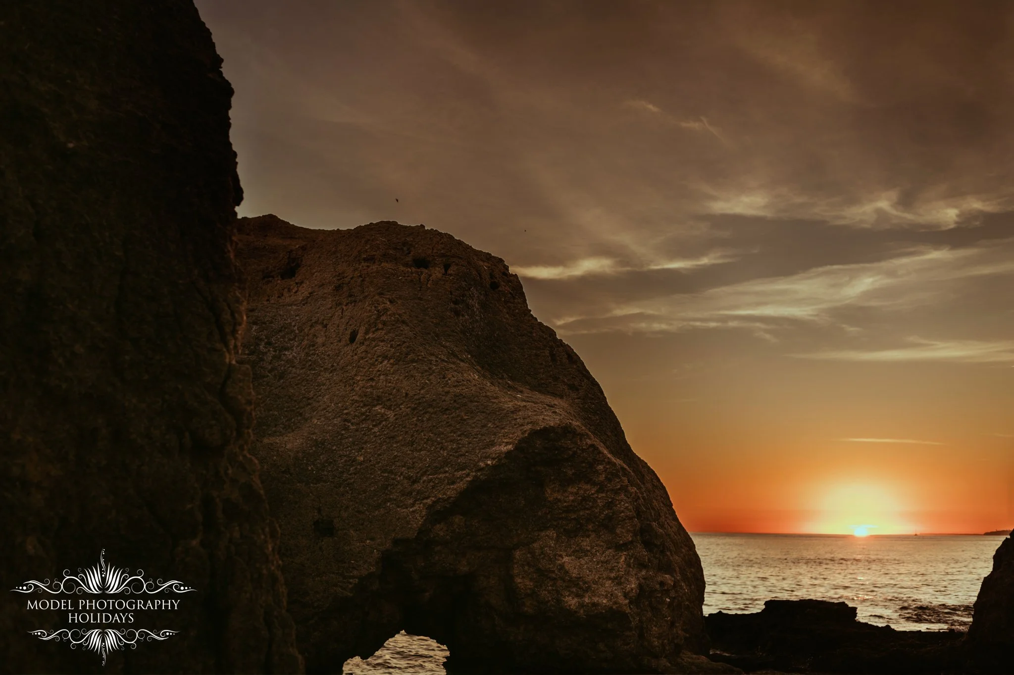 Sunset view through a rocky seaside landscape with large rocks in the foreground and an orange and yellow sky with clouds.