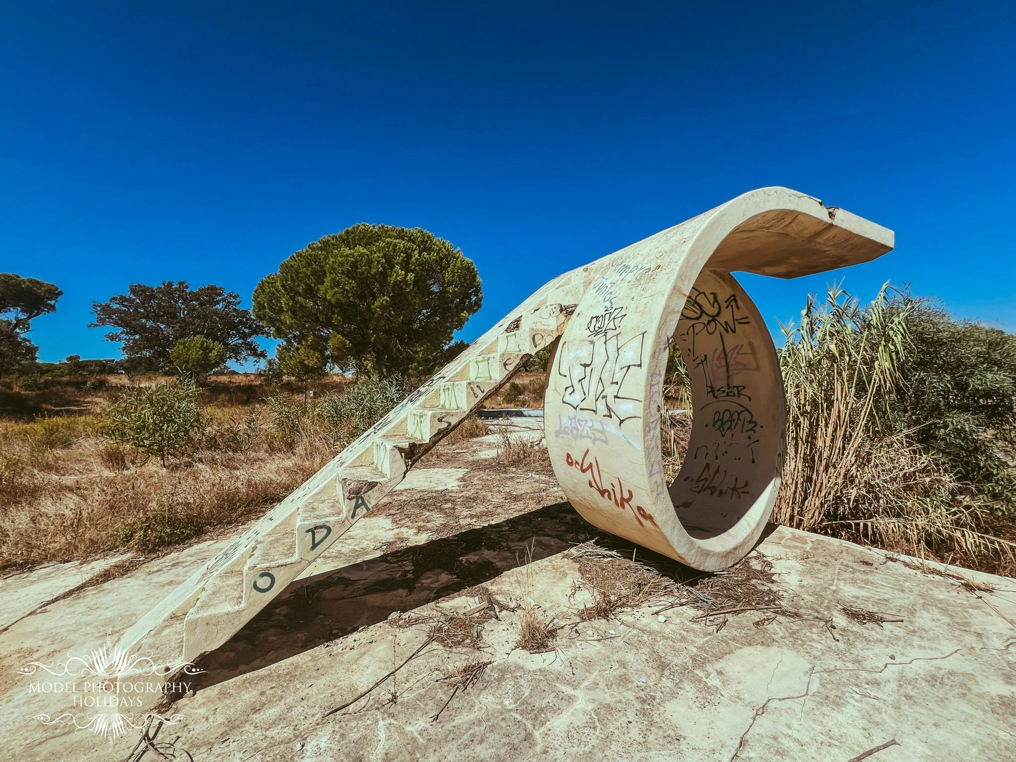 A large, broken concrete sculpture of a roll of toilet paper with graffiti and writing on it, situated outdoors in a dry natural landscape with trees and bushes under a clear blue sky.