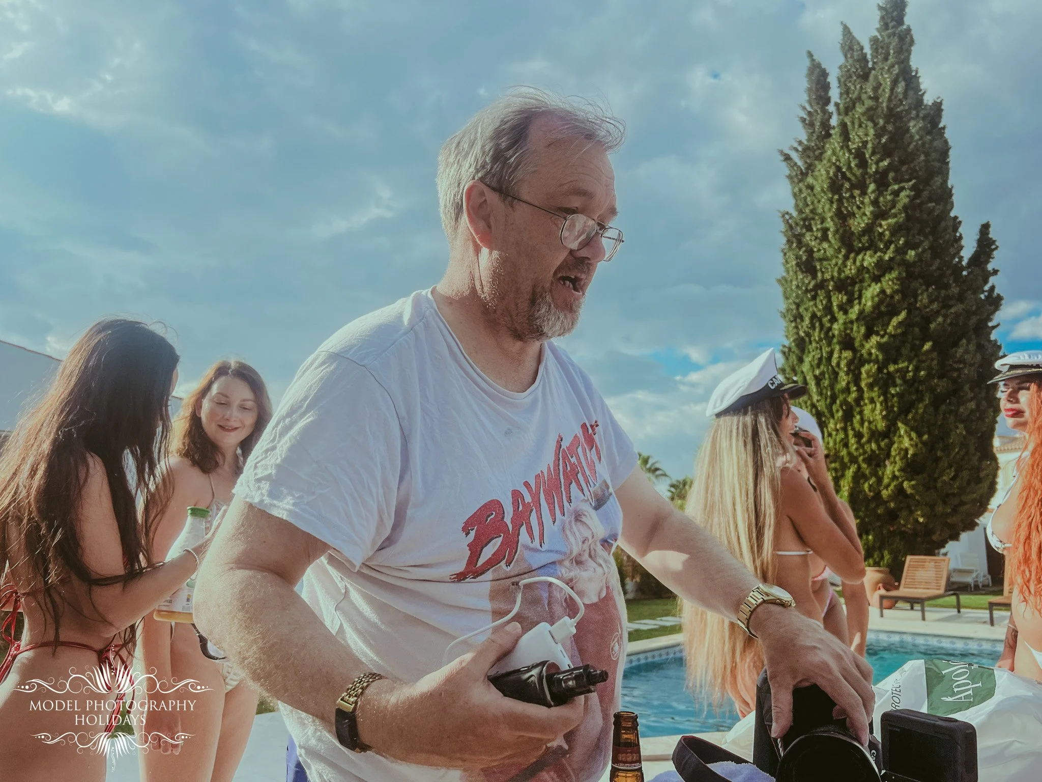 A group of women and a man gathered outdoors near a pool, with blue sky and clouds above, engaging in a social activity.