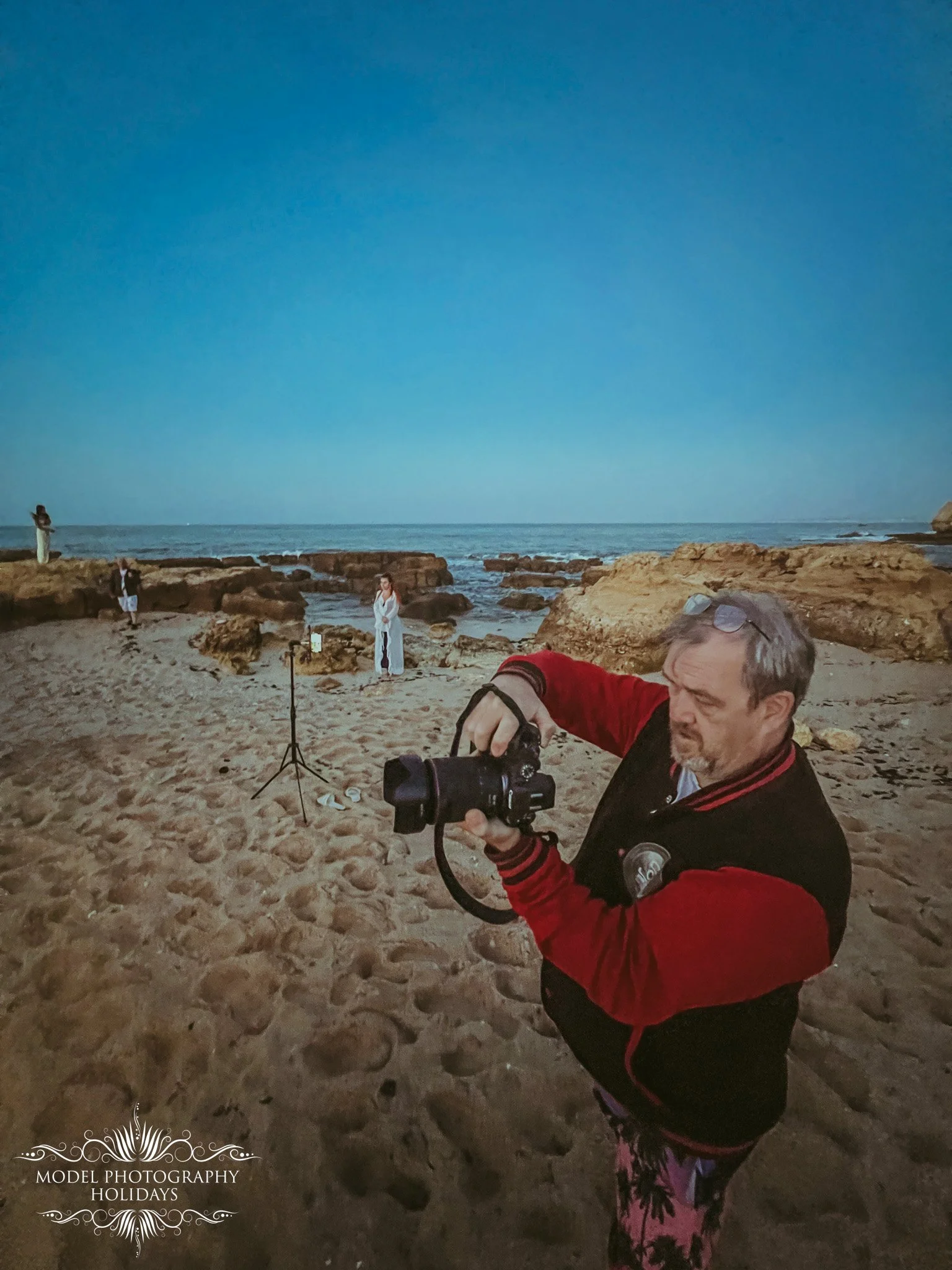 A man in a red and black jacket is looking at a camera in his hands on a sandy beach with rocky outcrops, during sunset or twilight. In the background, there are three women standing near rocks, with one holding a phone to take a photo, and others st