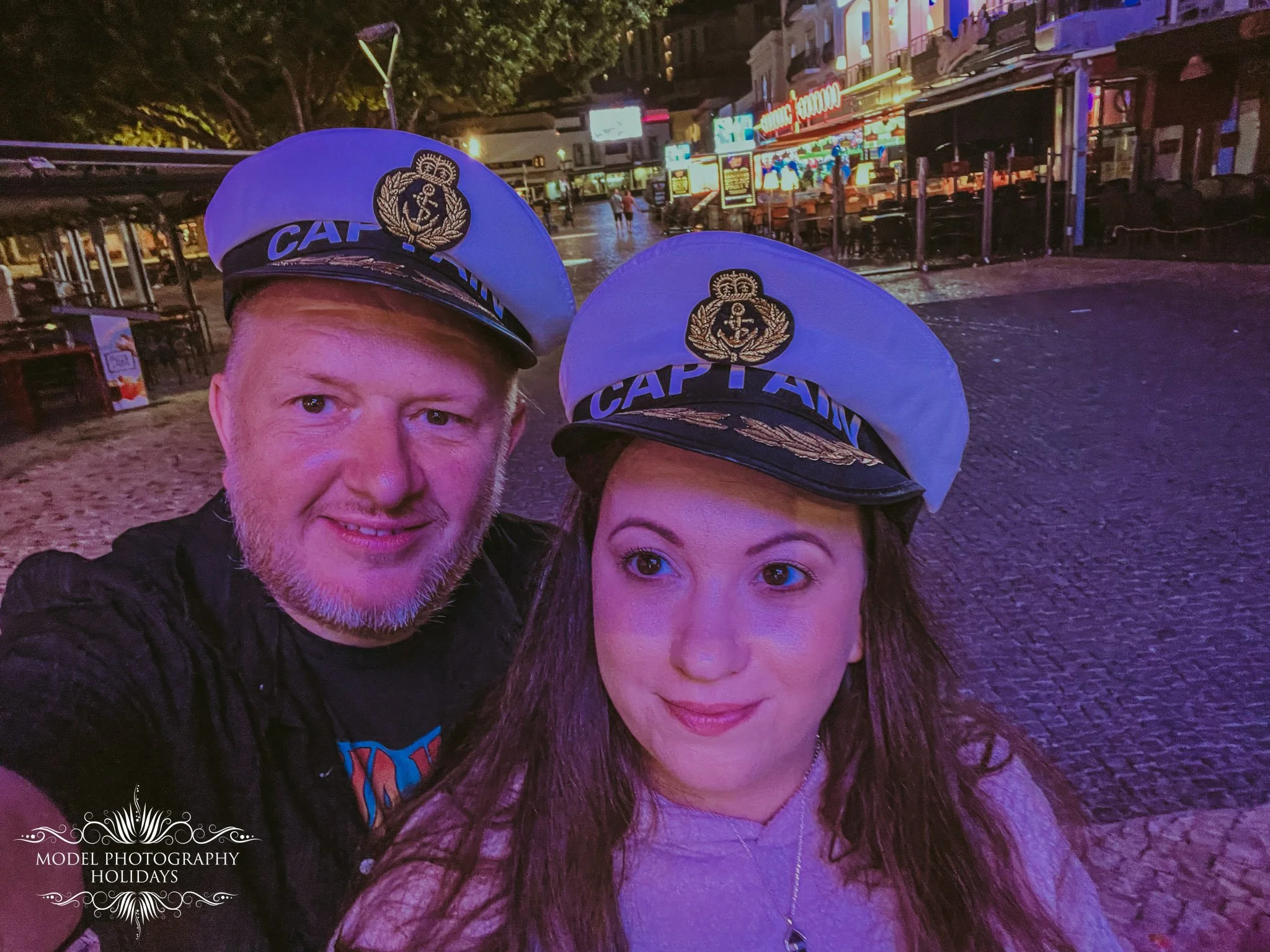A man and woman wearing white captain's hats with an anchor emblem taking a selfie in a lively, brightly lit street at night. The street has colorful neon signs and storefronts in the background.