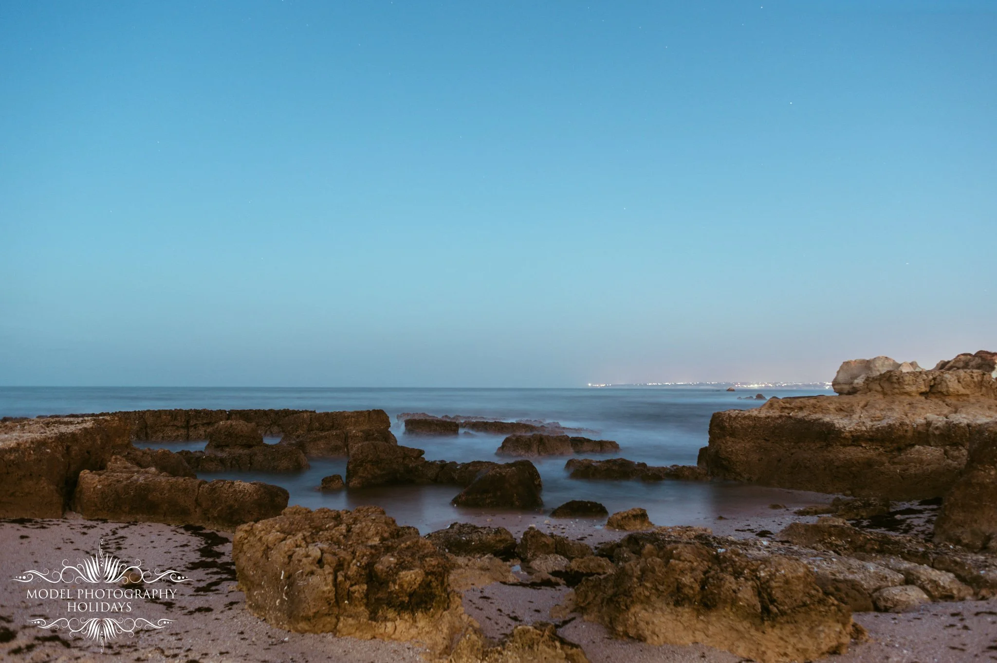 Nighttime view of a rocky beach with the ocean in the background, clear sky with faint stars, and city lights visible on the horizon.