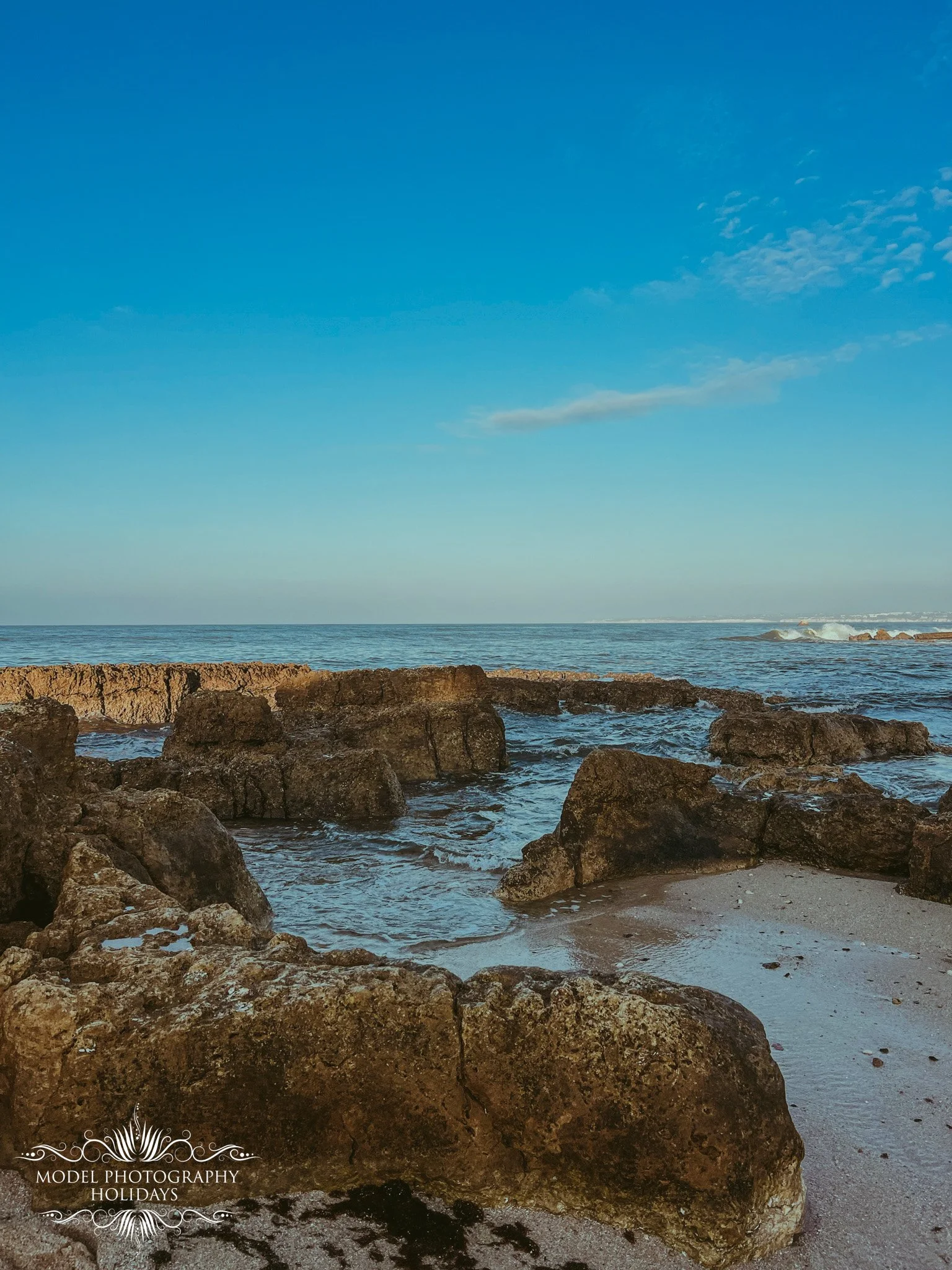 View of the ocean with rocks in the foreground, a clear blue sky, and some clouds near the horizon.