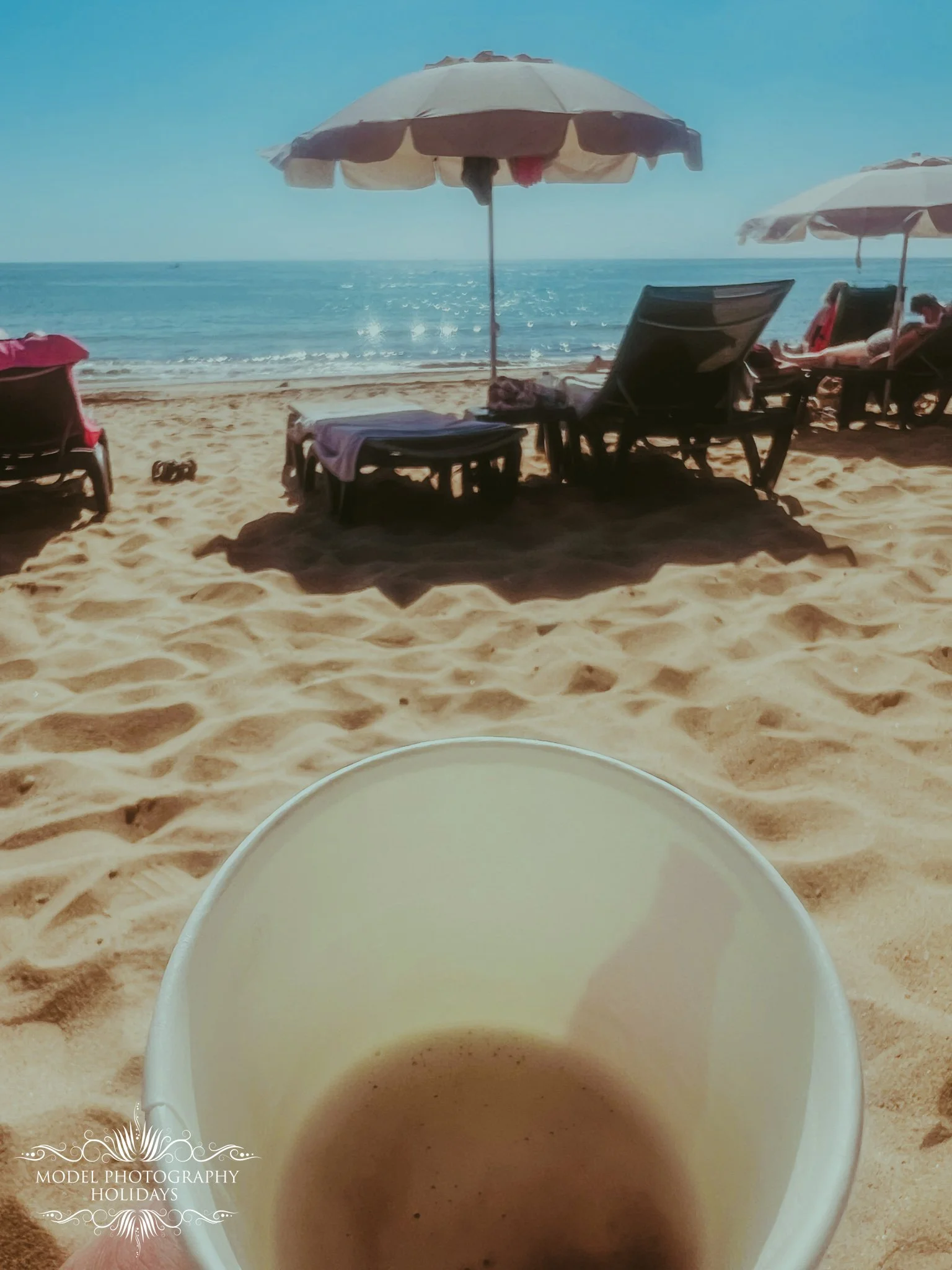 Close-up of a white coffee cup filled with a brown beverage in the foreground, with a sandy beach, beach chairs, umbrellas, and the ocean in the background on a sunny day.