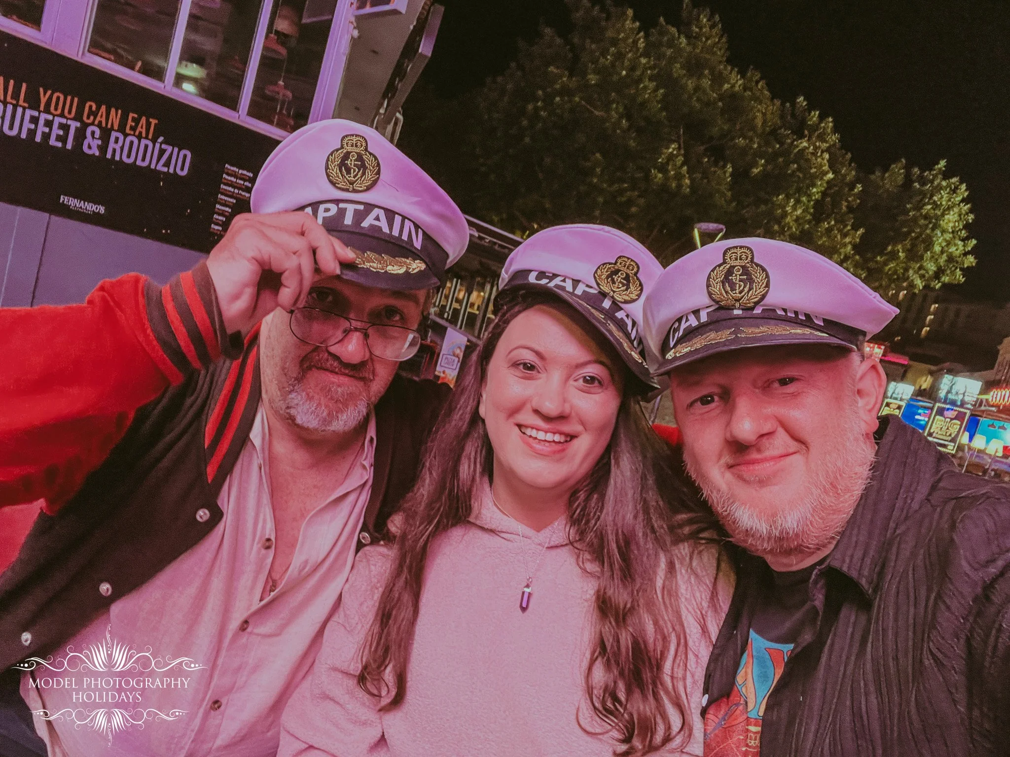 Three people wearing captain hats taking a selfie at an outdoor event during nighttime. Two men and a woman are smiling, with trees and a building in the background.