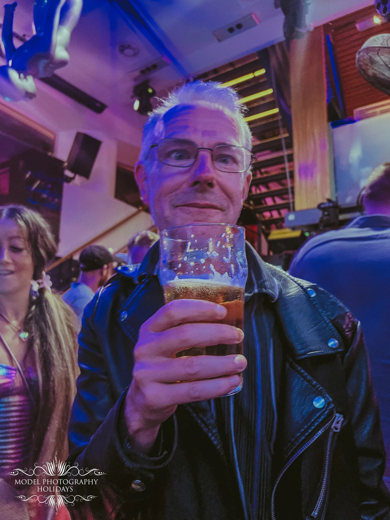 A man with gray hair, glasses, and a leather jacket holding a glass of beer at a social event with colorful lighting. There are other people in the background in a lively indoor setting.