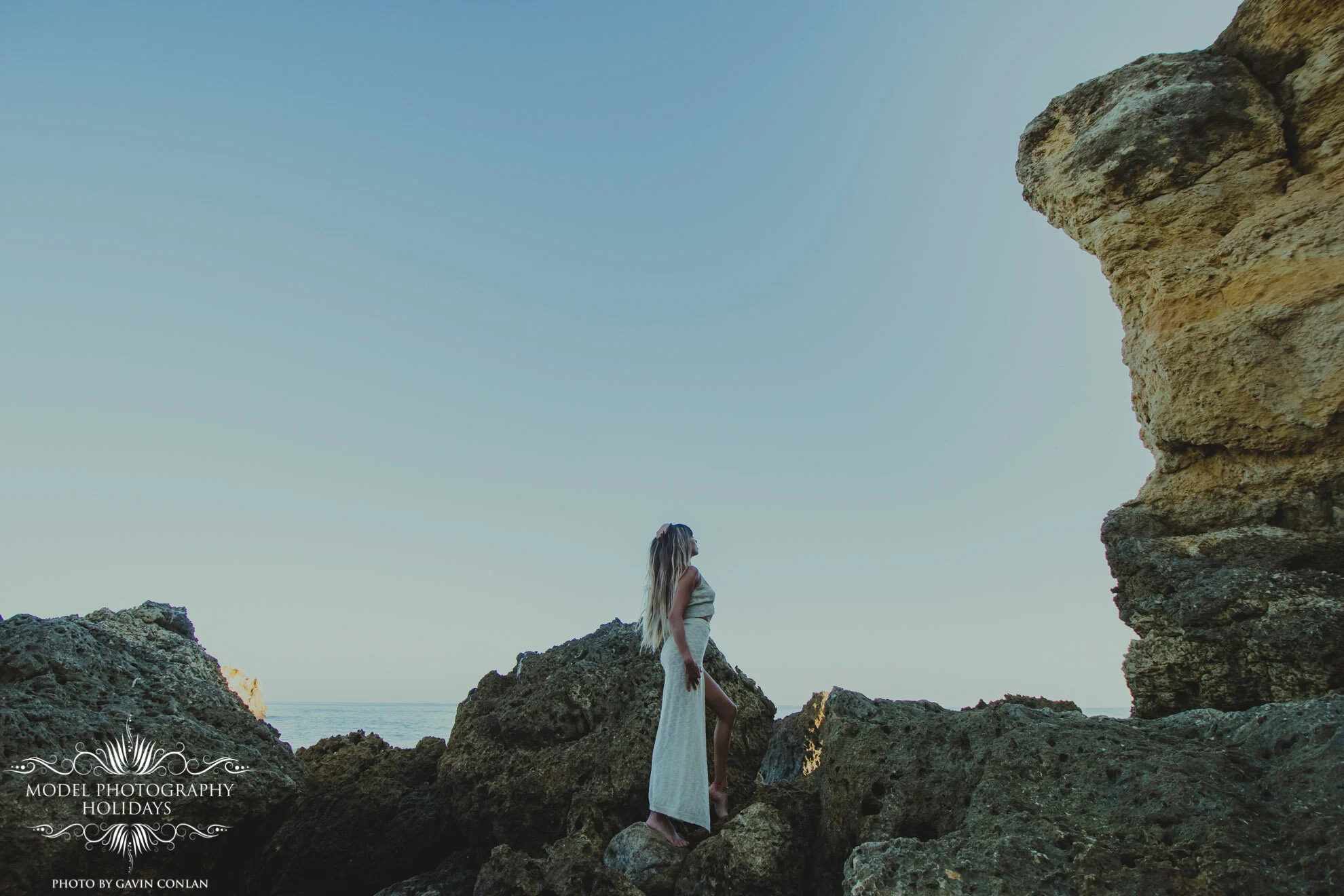 A woman in a long dress standing on large rocks by the sea under a clear blue sky.