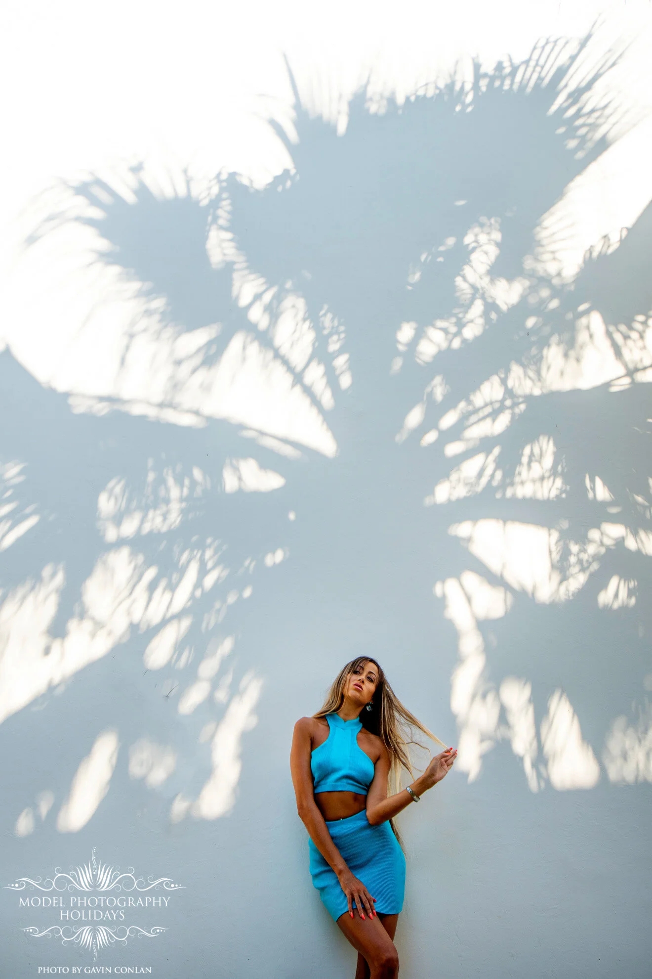 Woman in blue dress posing against a white wall with palm tree shadows.