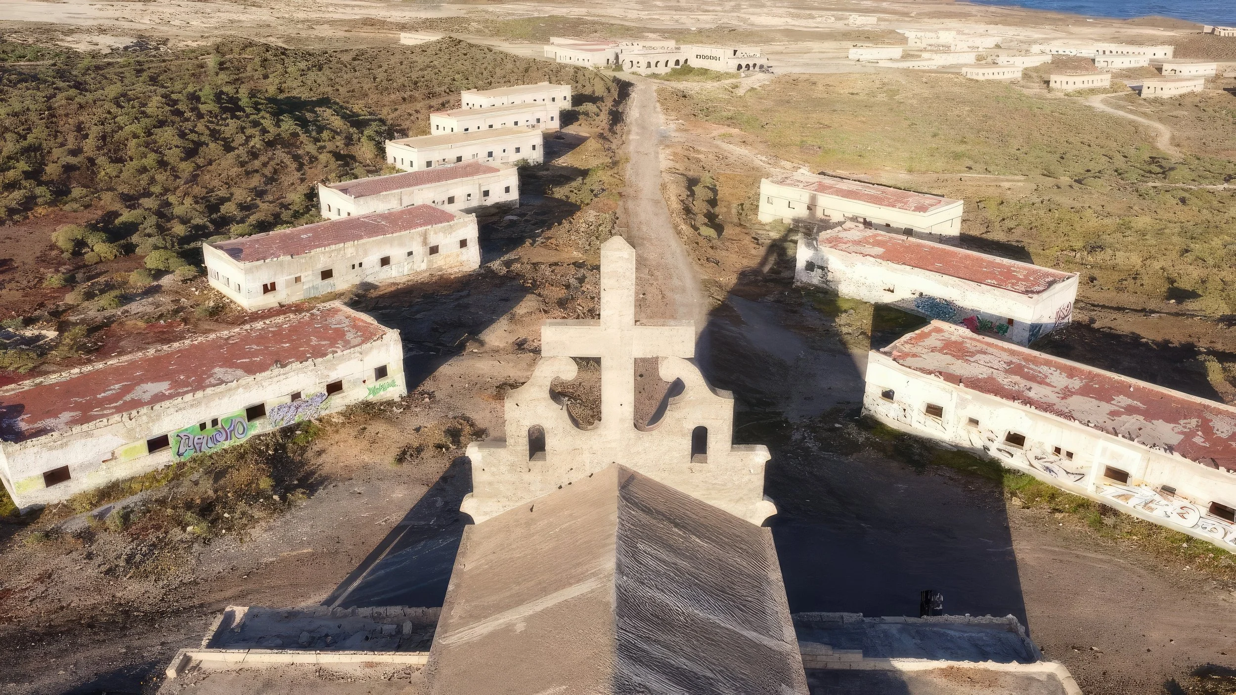 An aerial view of an abandoned building complex with multiple white, dilapidated structures and a large white cross-shaped monument in the foreground, surrounded by sparse vegetation and dirt roads.