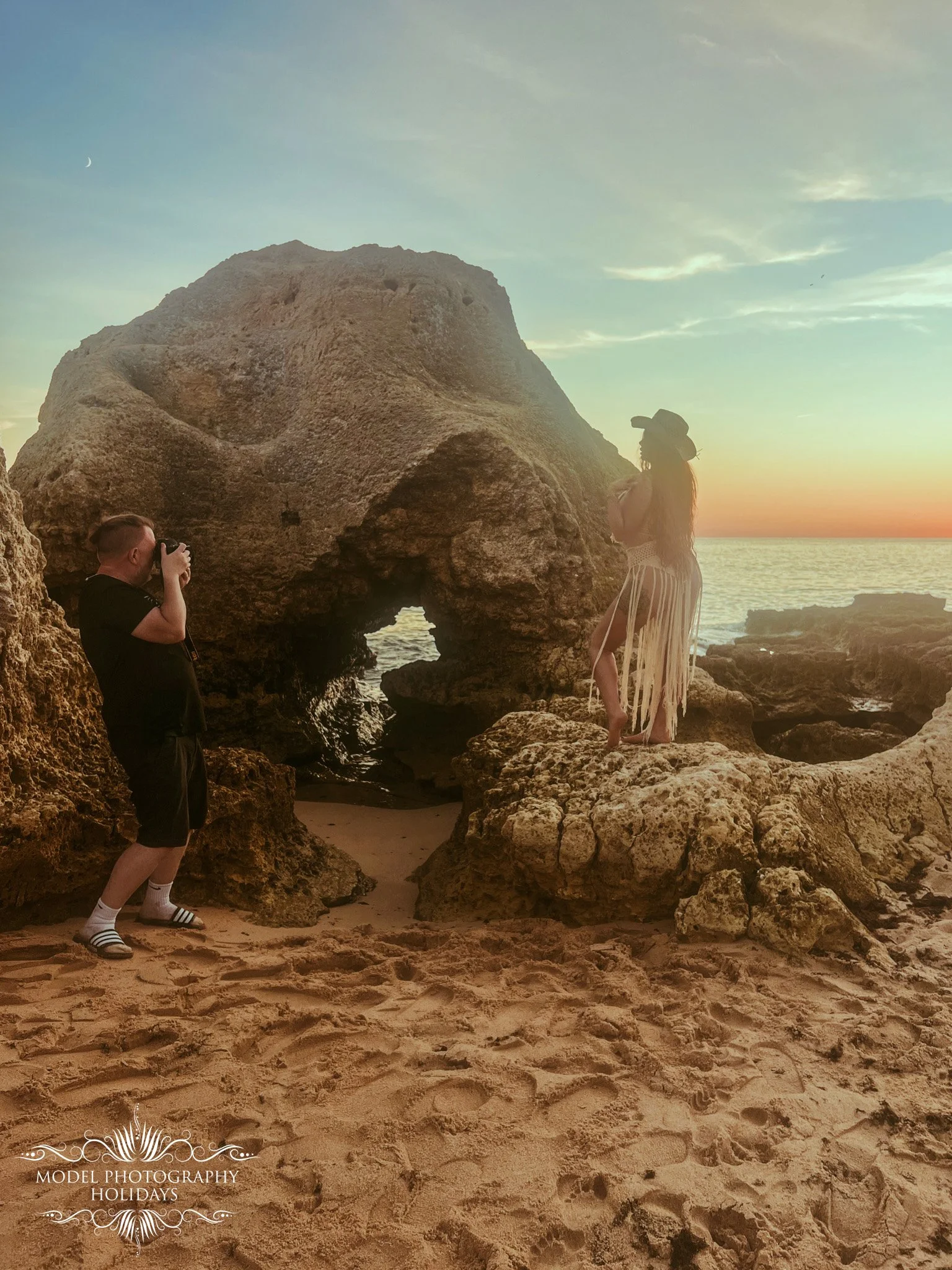 A person taking a photo of a woman posing on rocks near the ocean at sunset, with a large rock formation in the background and the sky with soft clouds and a crescent moon.