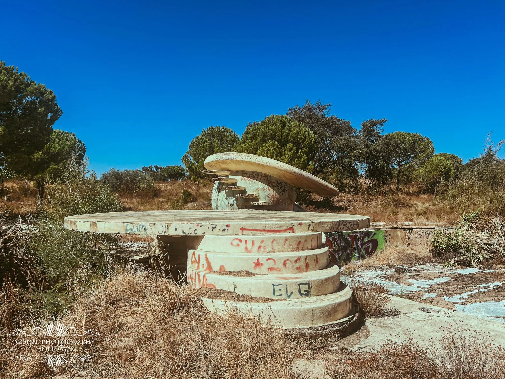 Stacked concrete rings with graffiti and spray paint in a dry outdoor area, surrounded by trees and grass under a clear blue sky.