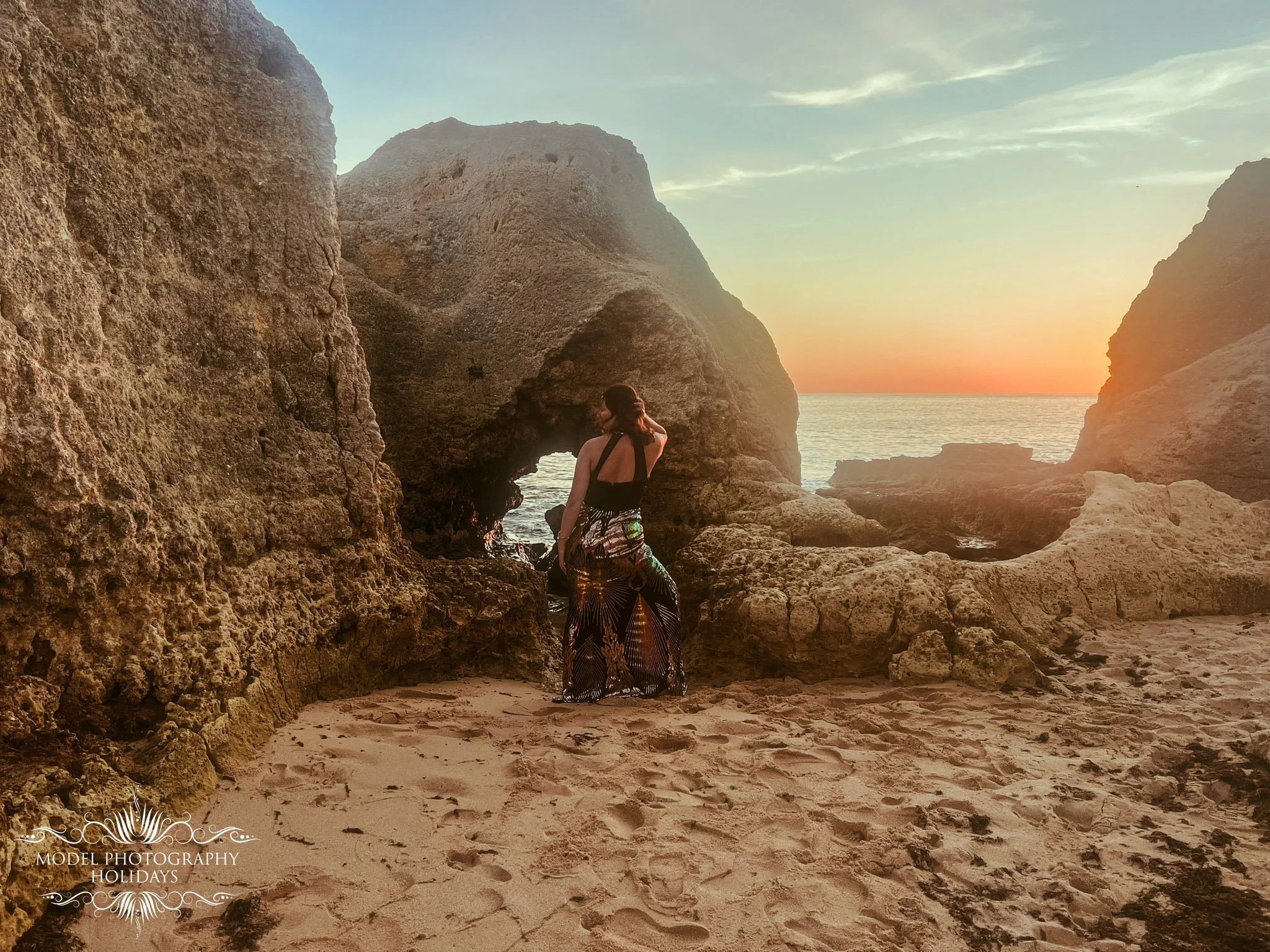 A woman in a patterned skirt and black top standing on a sandy beach surrounded by large rock formations at sunset, with ocean in the background.