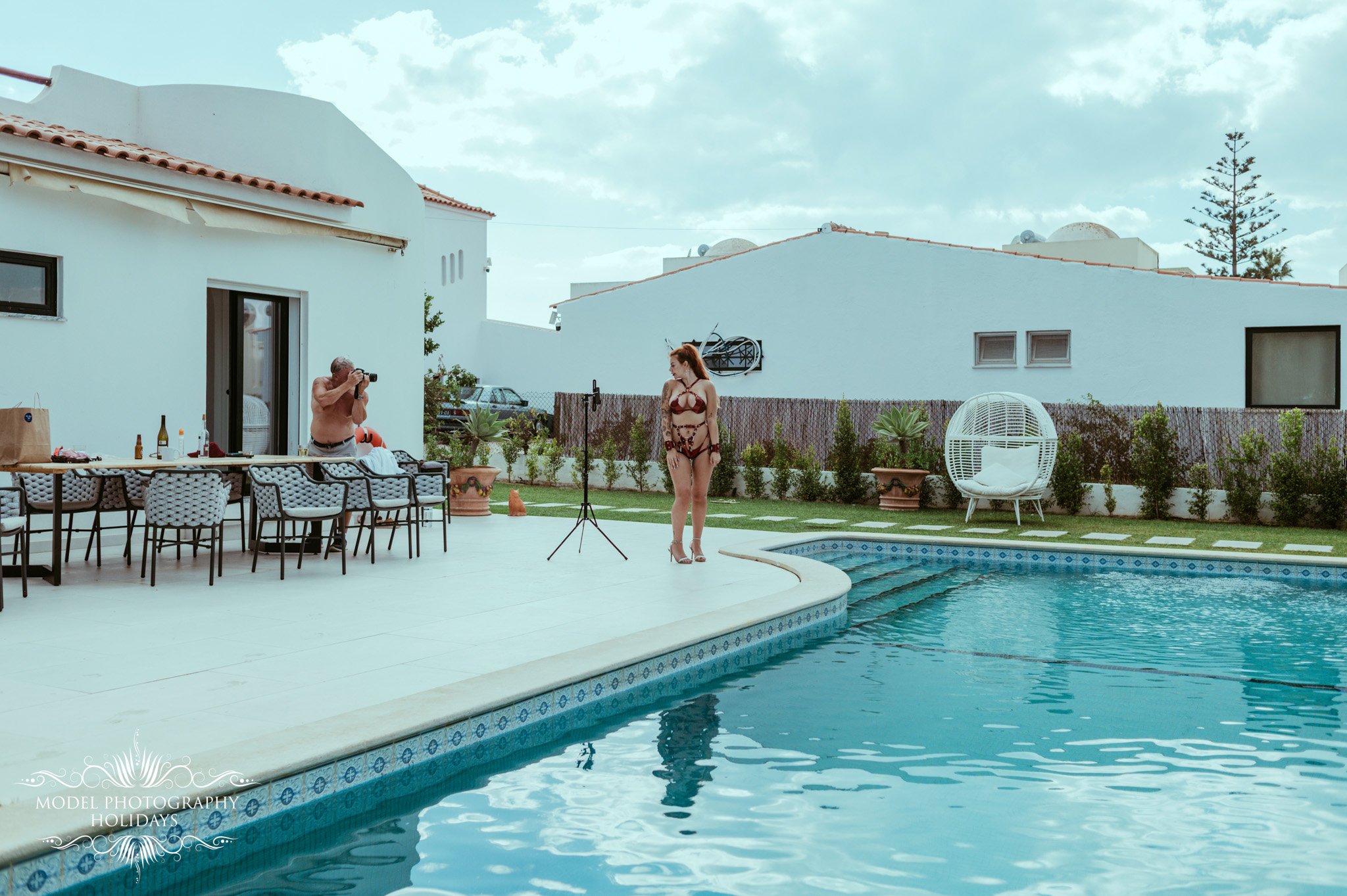 A woman in a bikini stands next to a camera on a tripod by a swimming pool, with a shirtless man taking photos nearby. There are outdoor furniture and potted plants, with modern white buildings and a cloudy sky in the background.