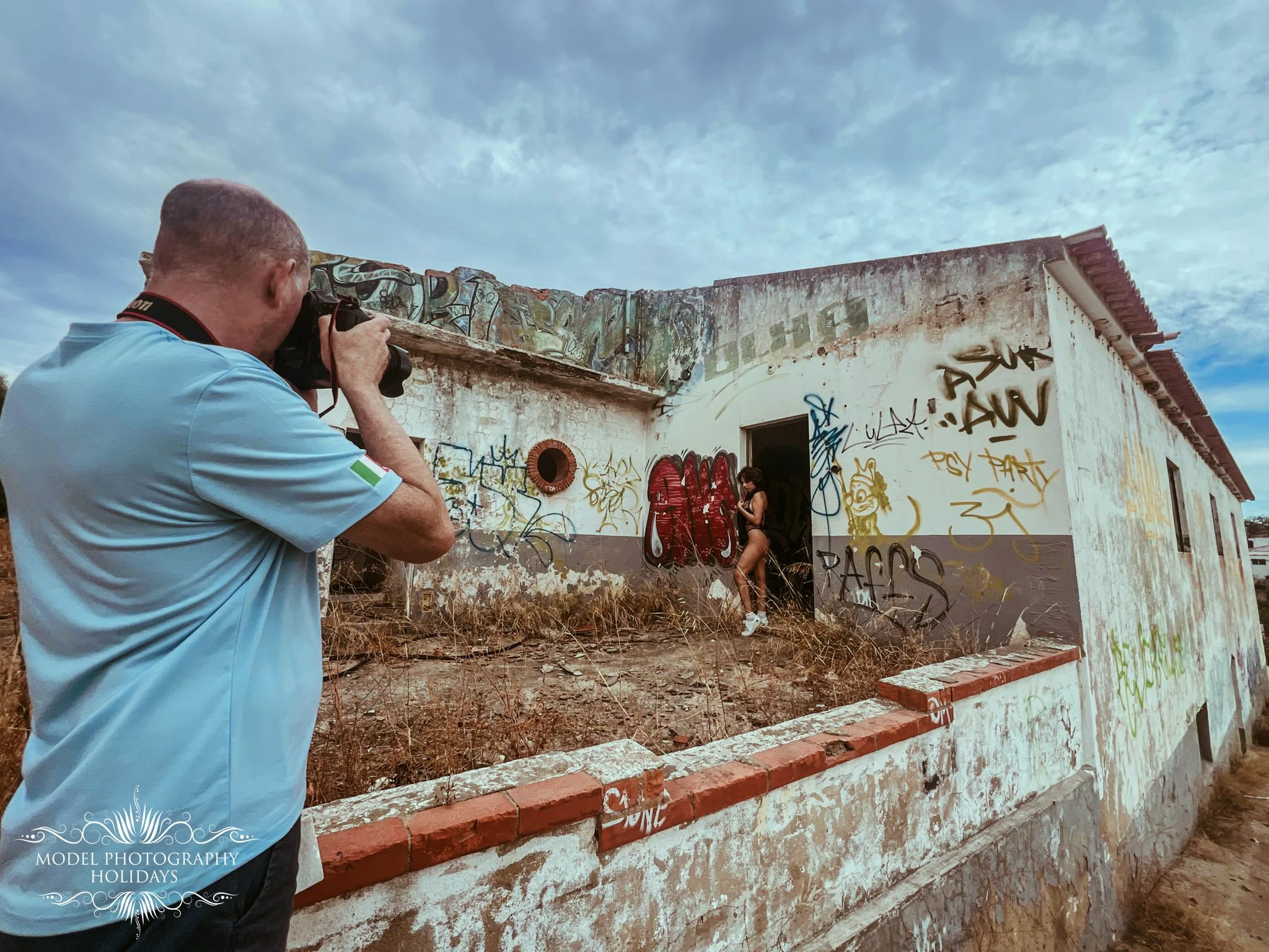 A man in a light blue shirt is taking a photograph of a graffitied building with a camera. The building has missing walls, exposed brick, and extensive graffiti art and tags. A woman in black clothing is standing near the building, and the sky is par