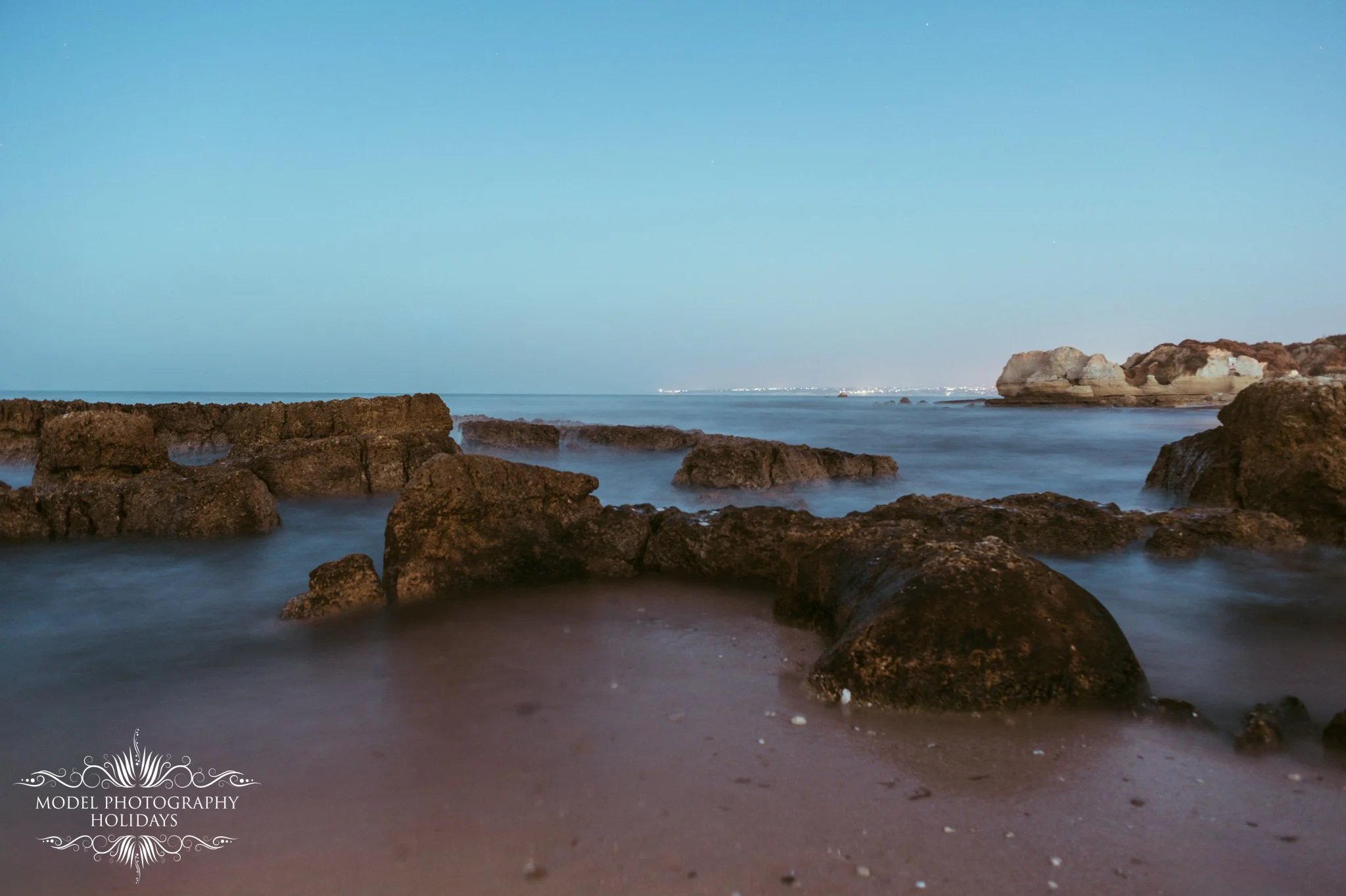 Nighttime view of a rocky beach with large rocks in the water and a calm sea, with a distant shoreline and city lights under a clear, starry sky.