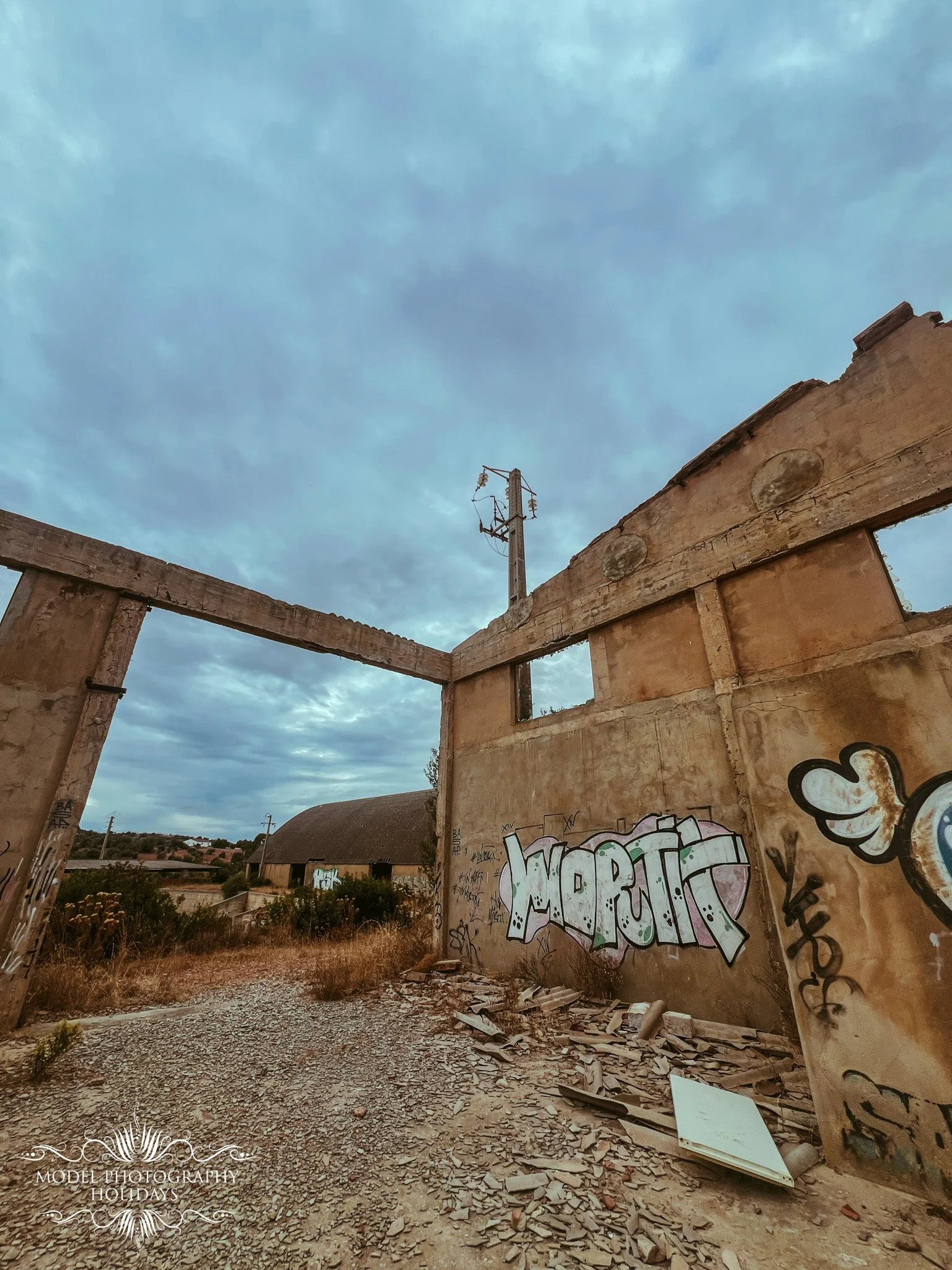 Abandoned building with graffiti on the walls, rubble and debris on the ground, and a view of a rural landscape with trees and houses in the background under a cloudy sky.