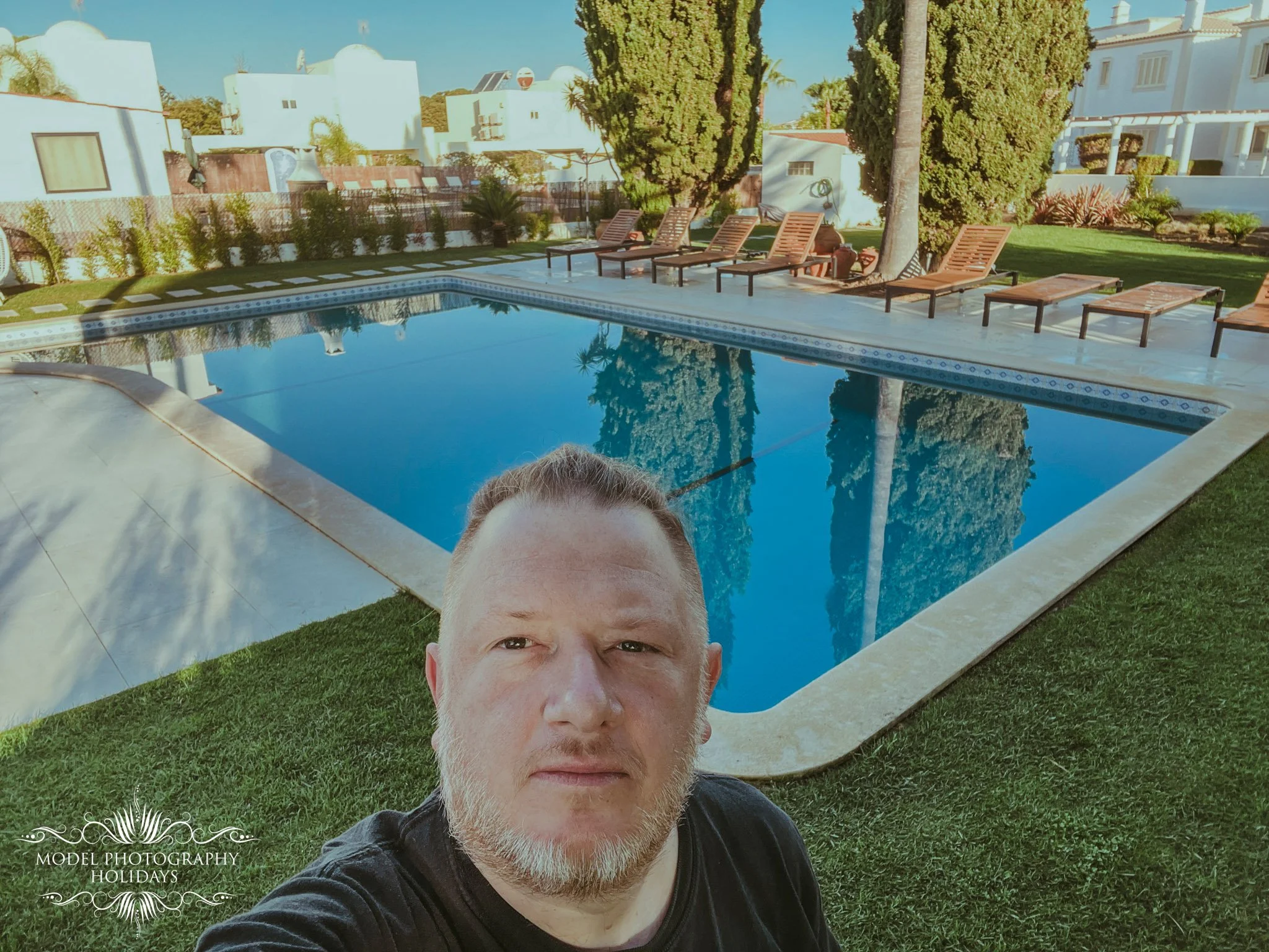 A man with a beard taking a selfie in front of a swimming pool in a backyard with green grass, tall trees, and white buildings in the background.