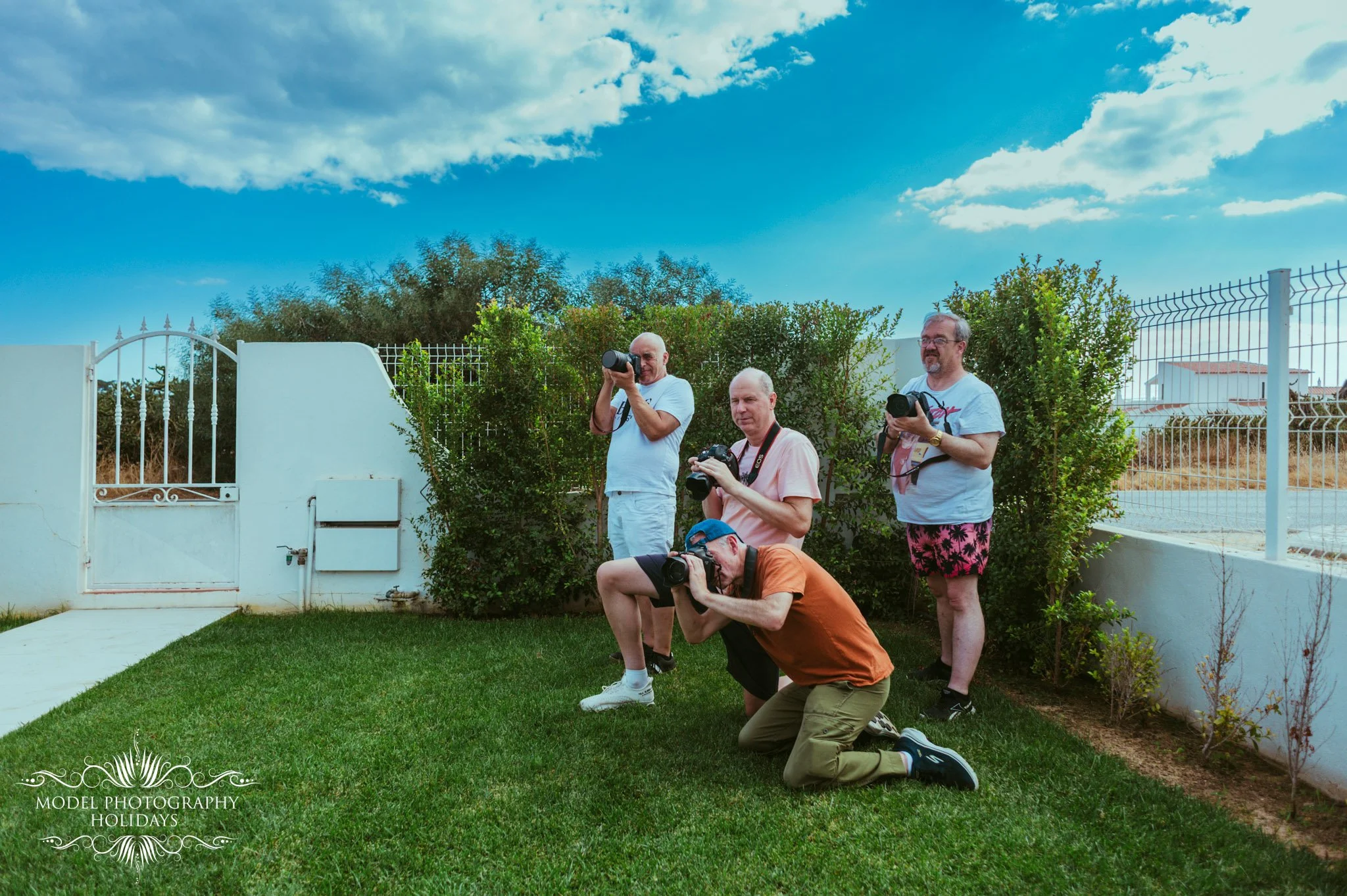 Four men taking photos outdoors in a residential area grass lawn, surrounded by bushes and a white fence, with a blue sky and clouds overhead.
