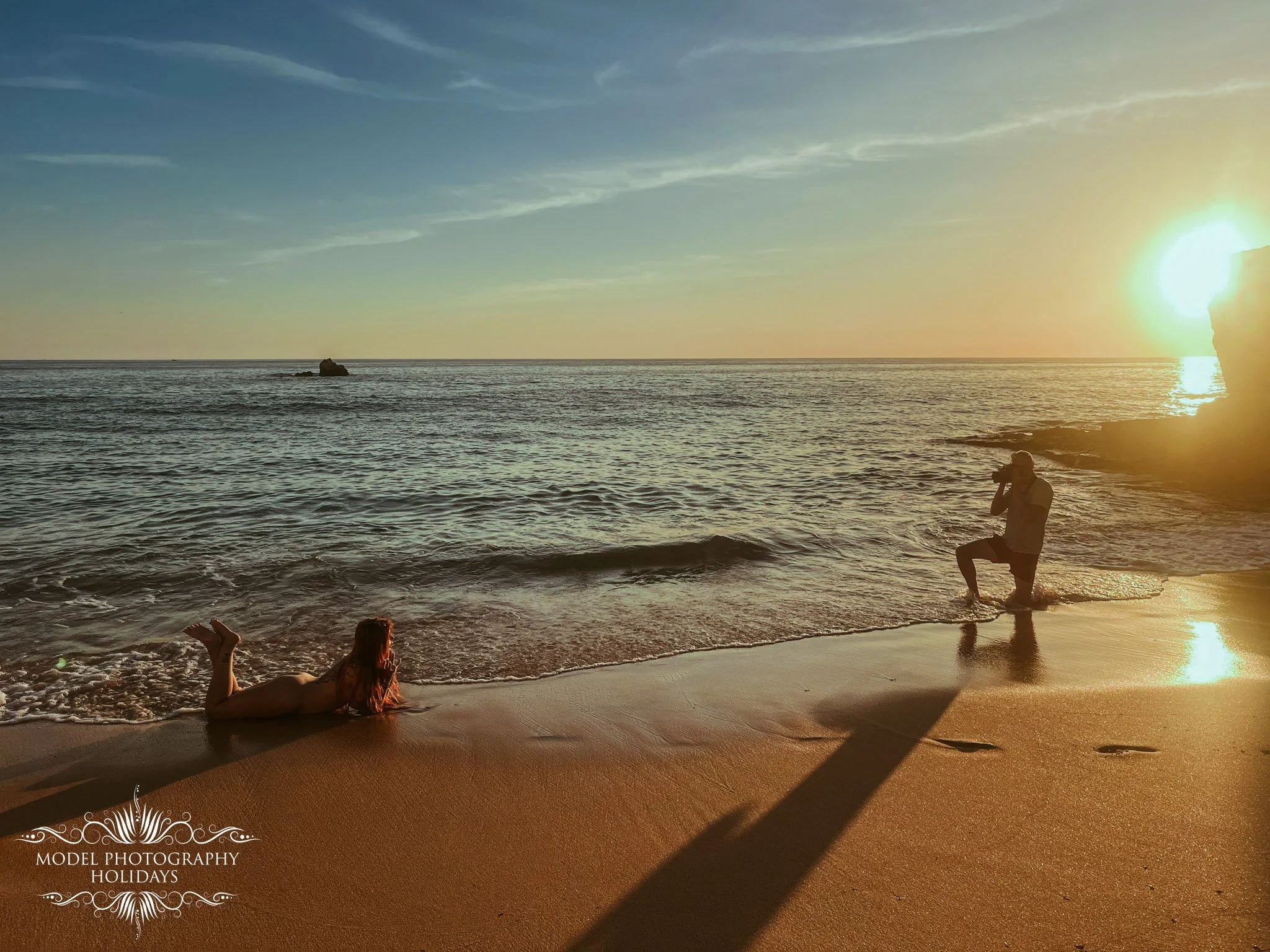 People relaxing on a beach at sunset, with one person lying on the sand and others near the water, the sky has wispy clouds and the sun is setting on the horizon.