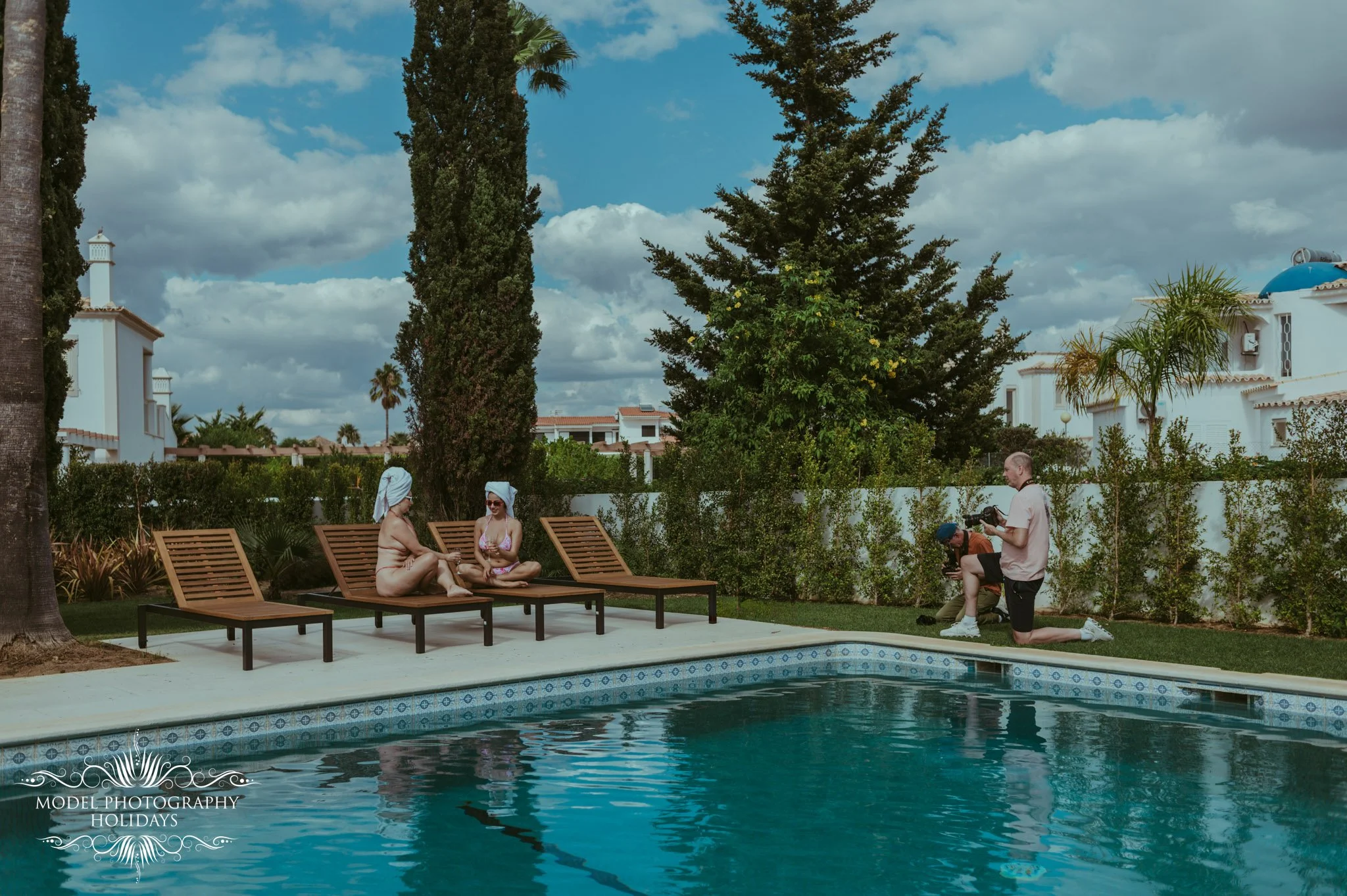 Two women in swimsuits and towels on their heads sit on lounge chairs by a swimming pool, with a man kneeling nearby taking a photograph of them. There are trees and white buildings with blue accents in the background under a partly cloudy sky.