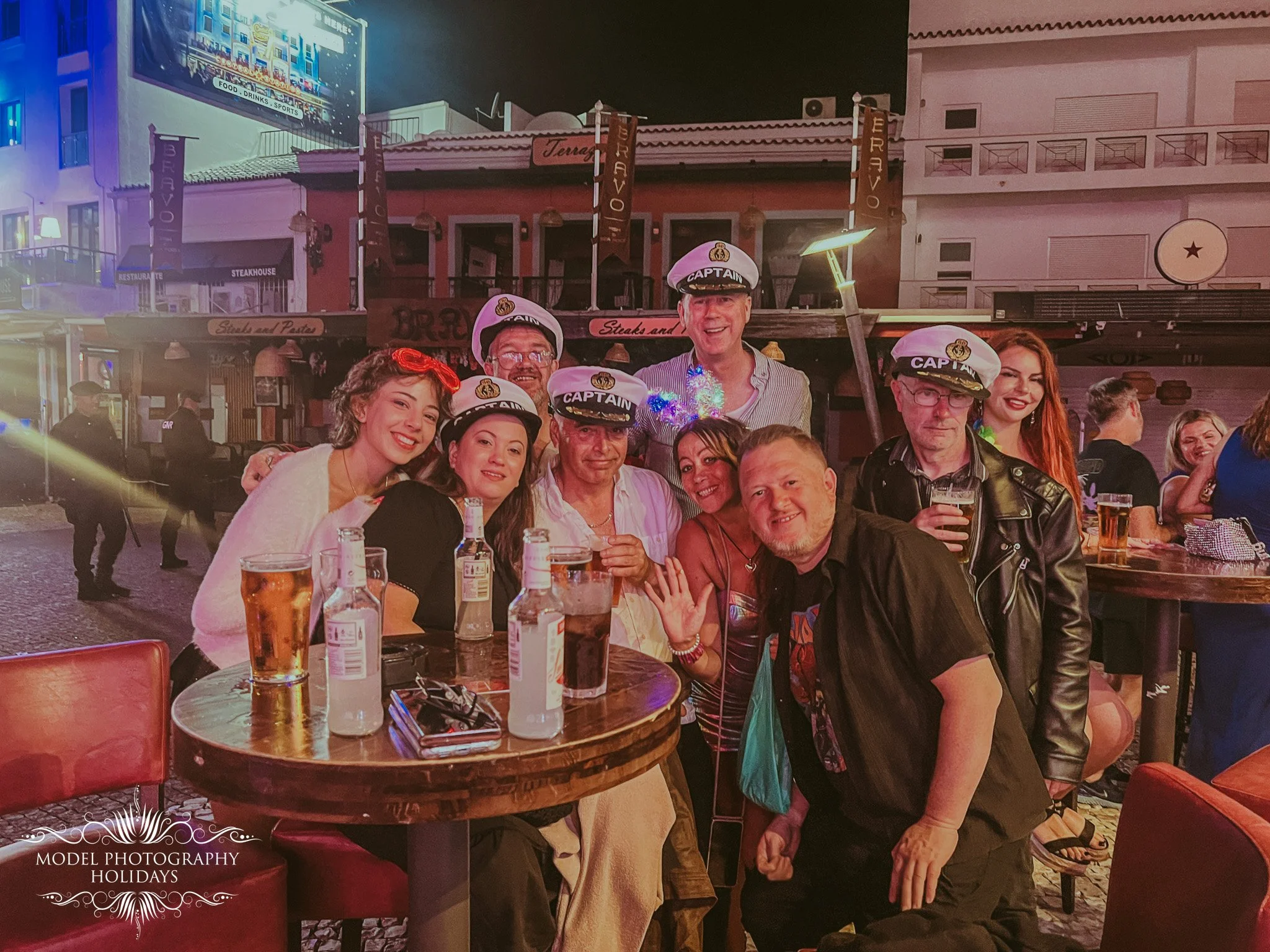 Group of people at an outdoor restaurant or bar in a lively nighttime setting. Several individuals are wearing captain hats, and there are drinks on the table. The background shows buildings and signage, indicating an energetic, social atmosphere.
