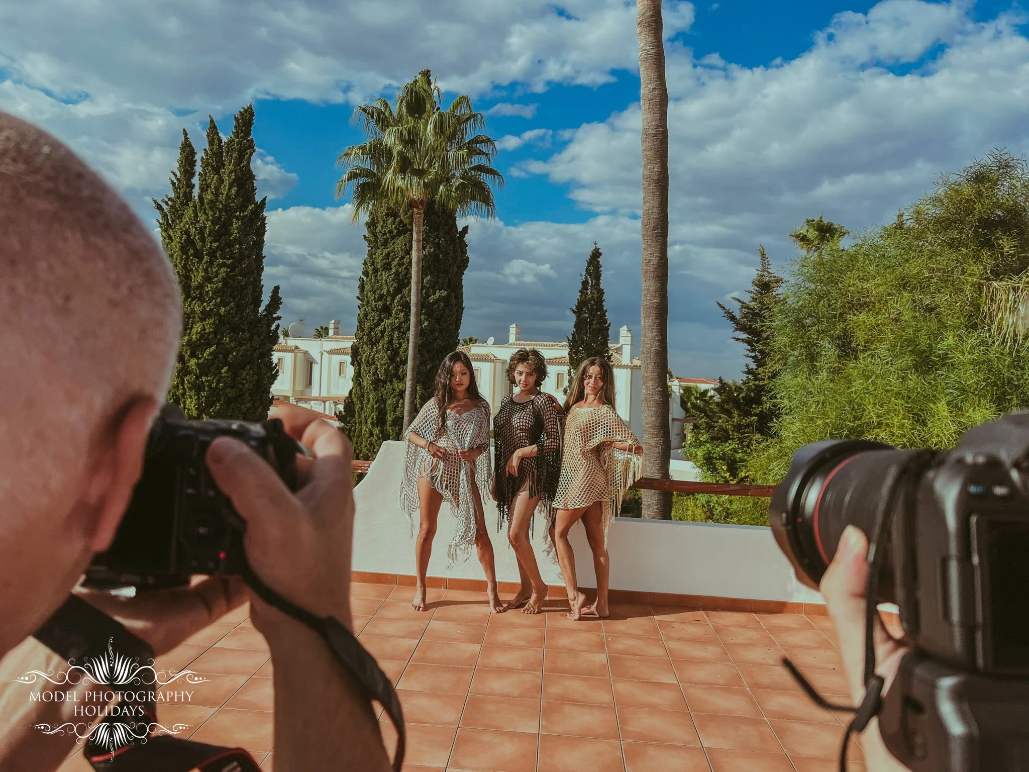 Three women posing outdoors on a sunny day with palm trees and greenery behind them, viewed through the camera and photographer's hands in the foreground.