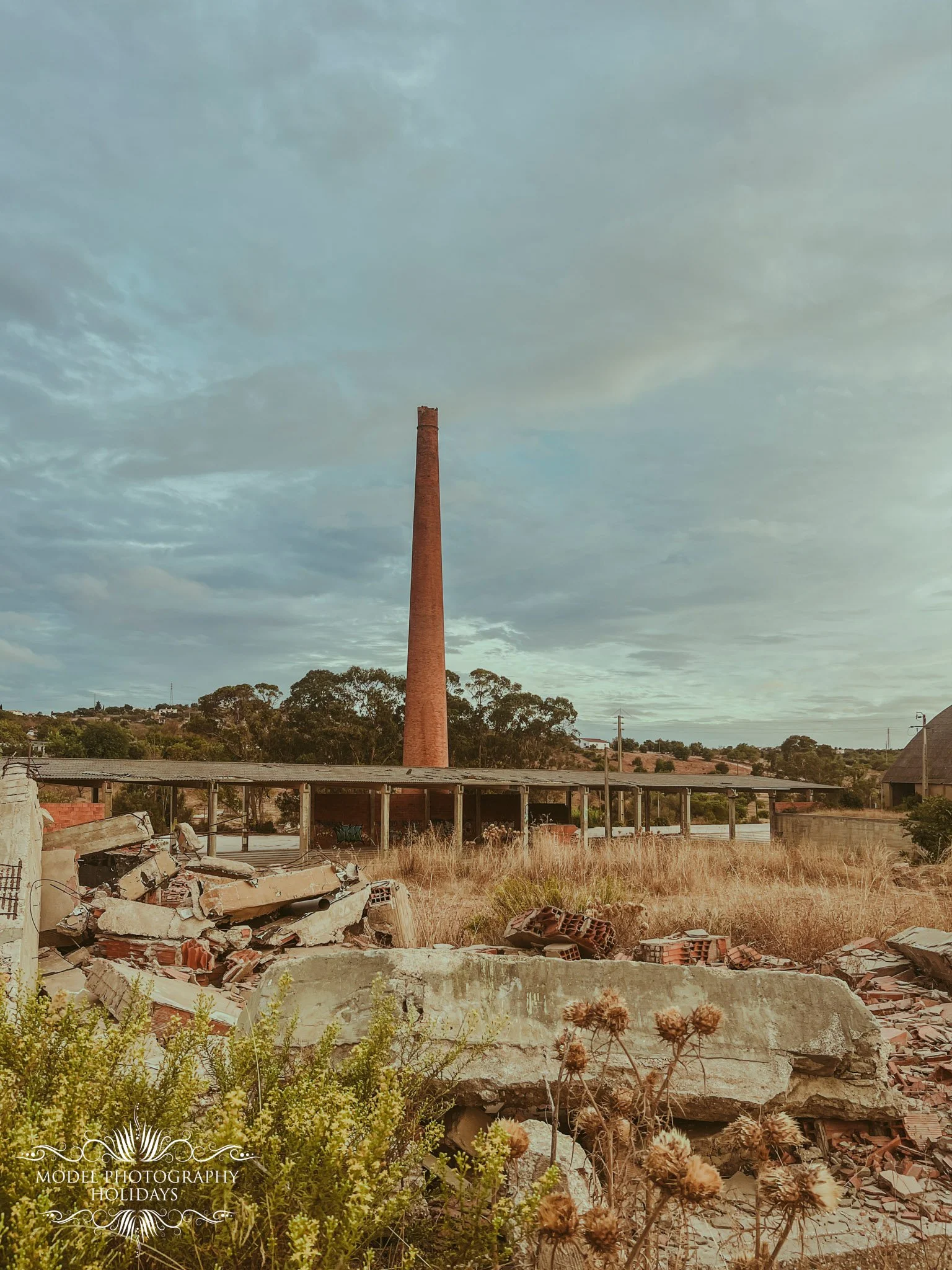 An old, abandoned building with a tall brick smokestack in the background, debris and rubble in the foreground, and overgrown vegetation; cloudy sky above.