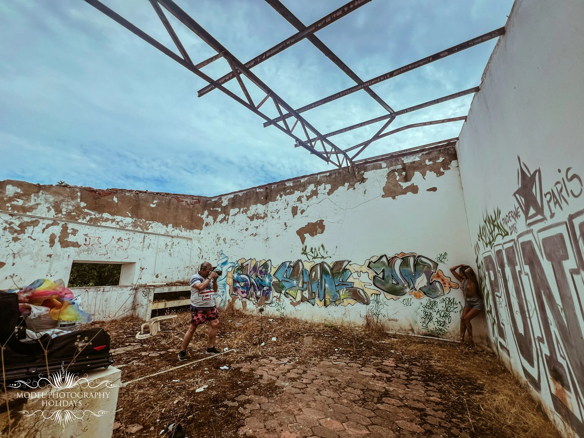 An outdoor scene with graffiti-covered walls, two people, one taking a photo and one standing, in a partially collapsed building with a metal structure overhead and a cloudy sky.