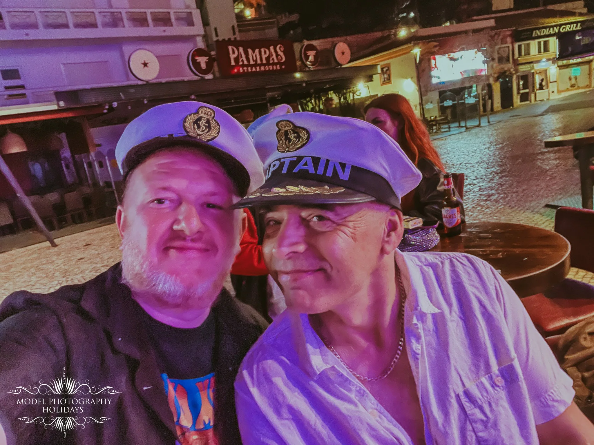 Two men wearing white Navy captain hats with tattoos on one man's arm, taking a selfie at night at an outdoor dining area. In the background, a woman with long reddish hair and a bottle of beer is sitting near a restaurant with signs for 'Pampas' and