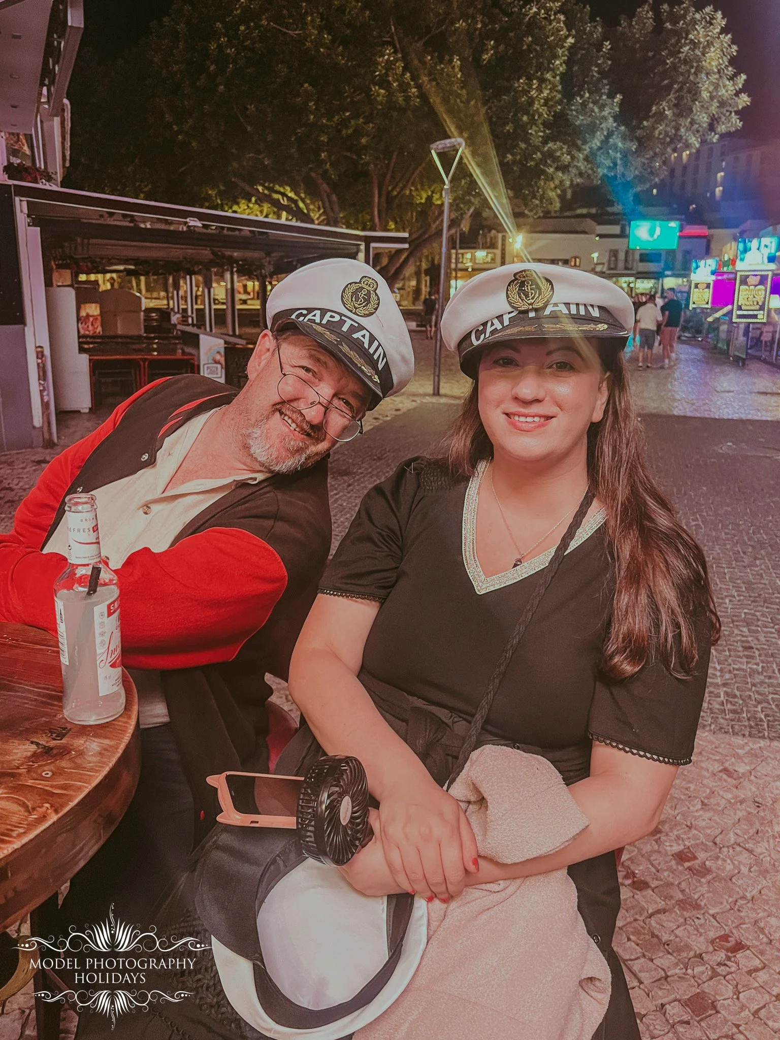 Two people wearing captain hats with maritime insignia pose for a photo outdoors at night, smiling. One, a man, wears glasses and a red and beige jacket, and the woman, holding a beige towel on her lap, has long brown hair and is dressed in a black t