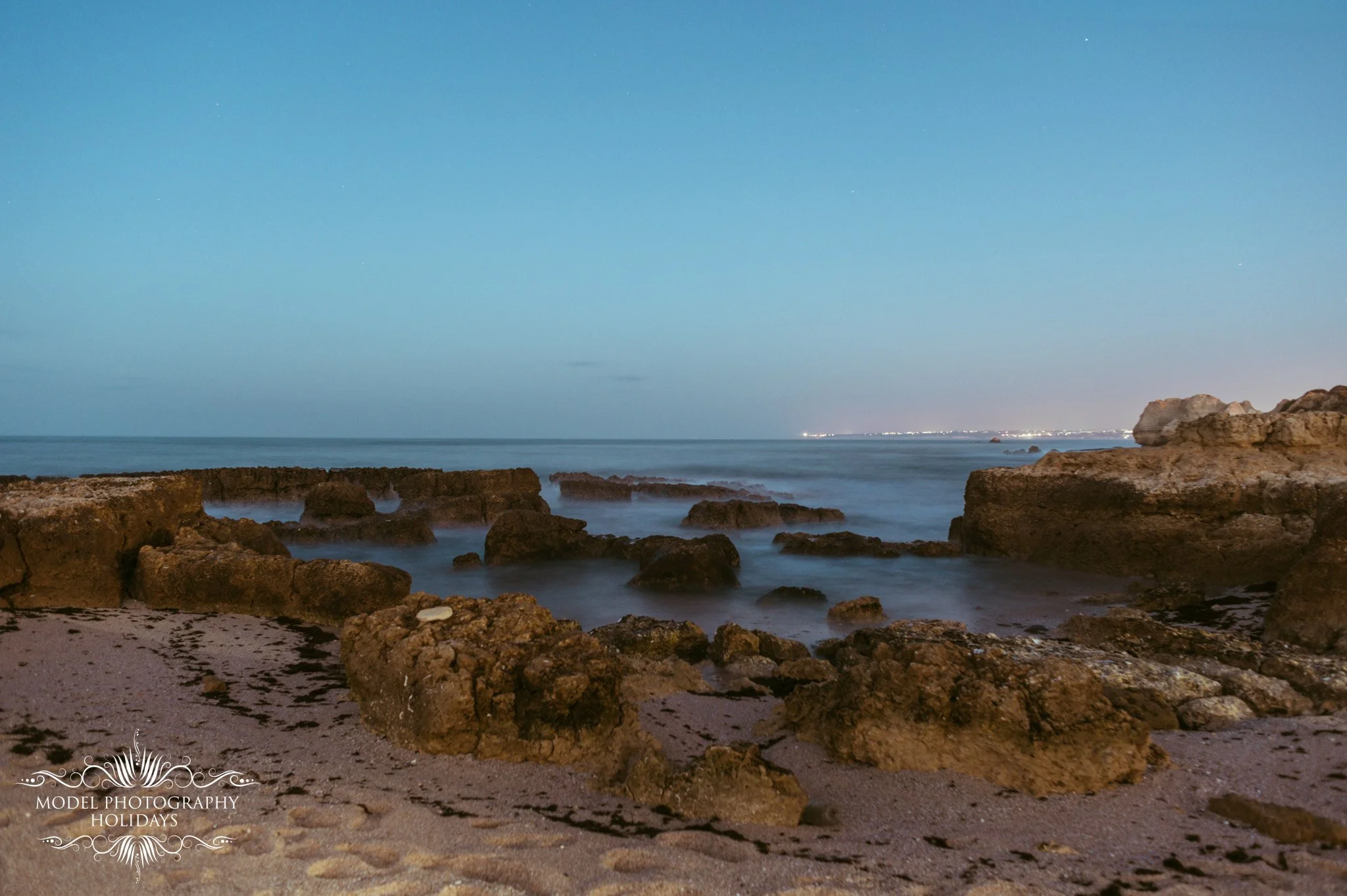 Nighttime view of the ocean with rocks in the foreground and city lights in the distance under a clear sky with stars.