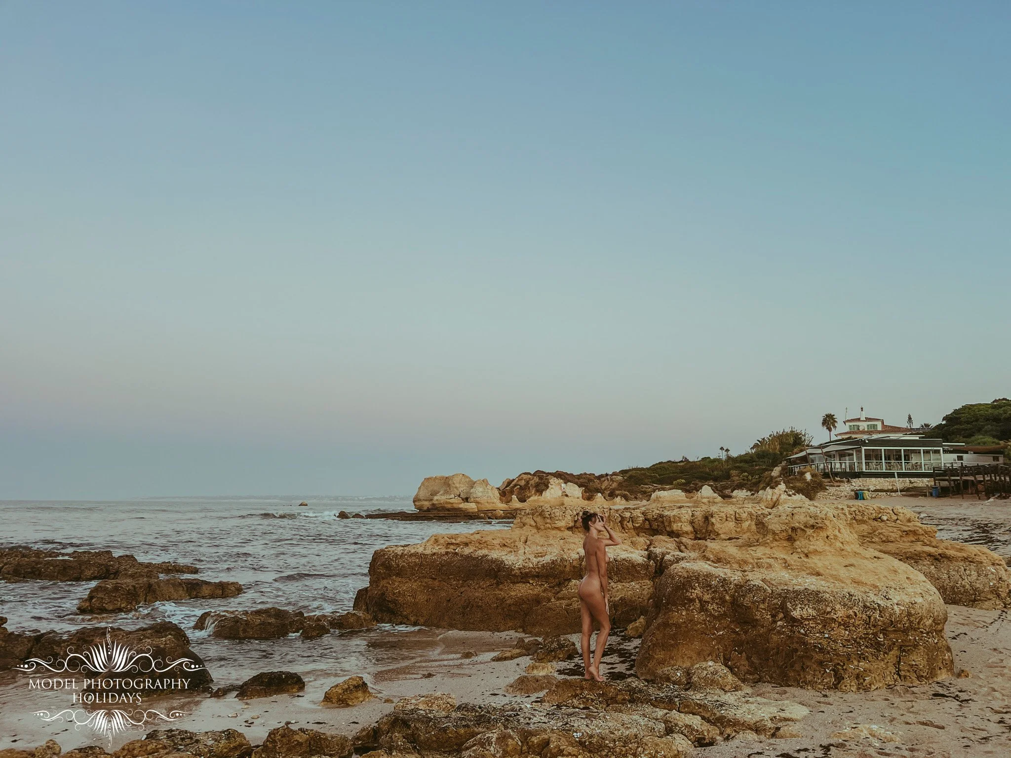 A woman standing nude on a rocky beach during sunset, with ocean waves and houses on a hill in the background.