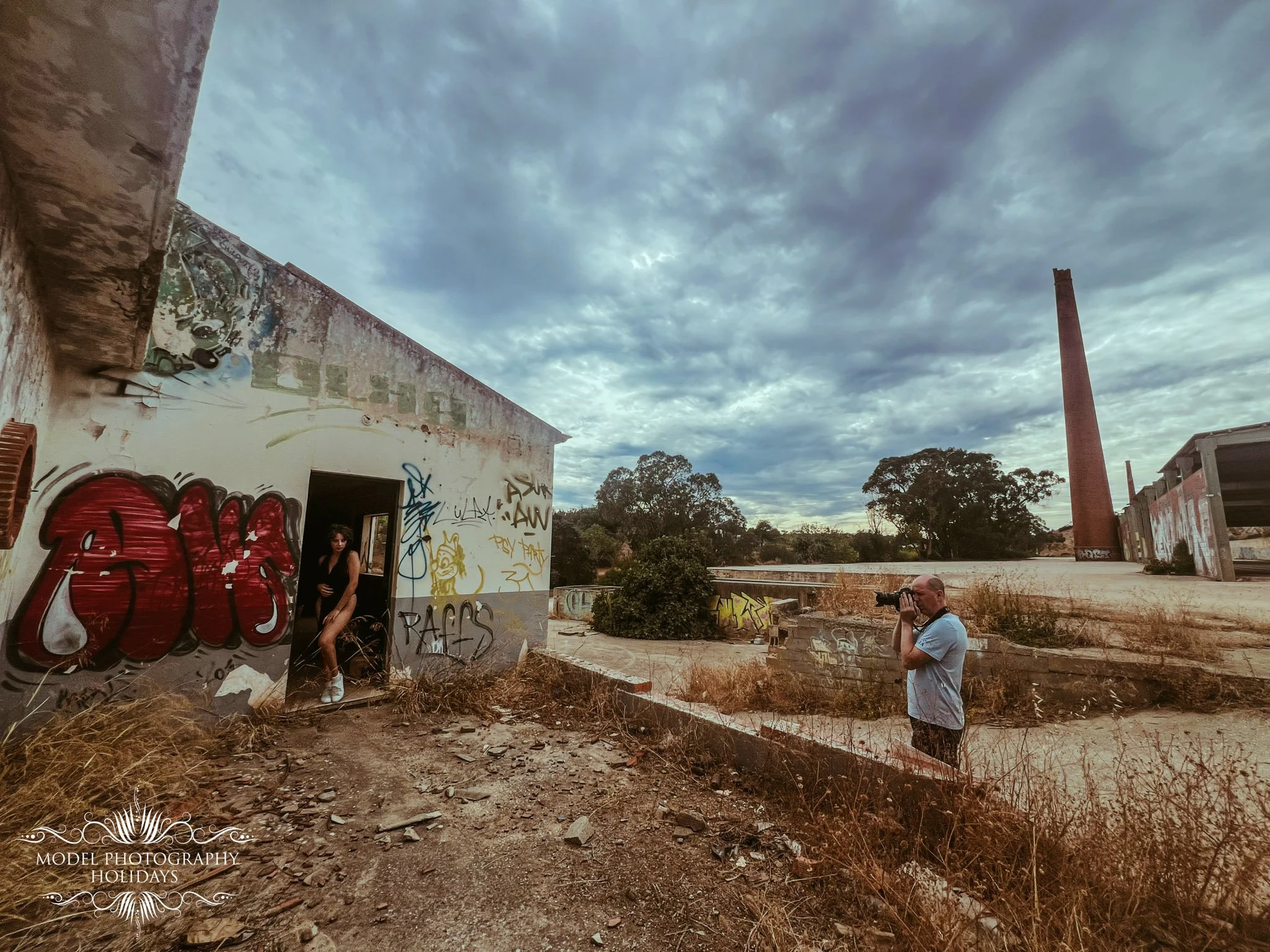 An abandoned building with graffiti on the walls, a large chimney, and a cloudy sky sky with trees in the background. There are two people, one man with a camera and a woman standing in a doorway.