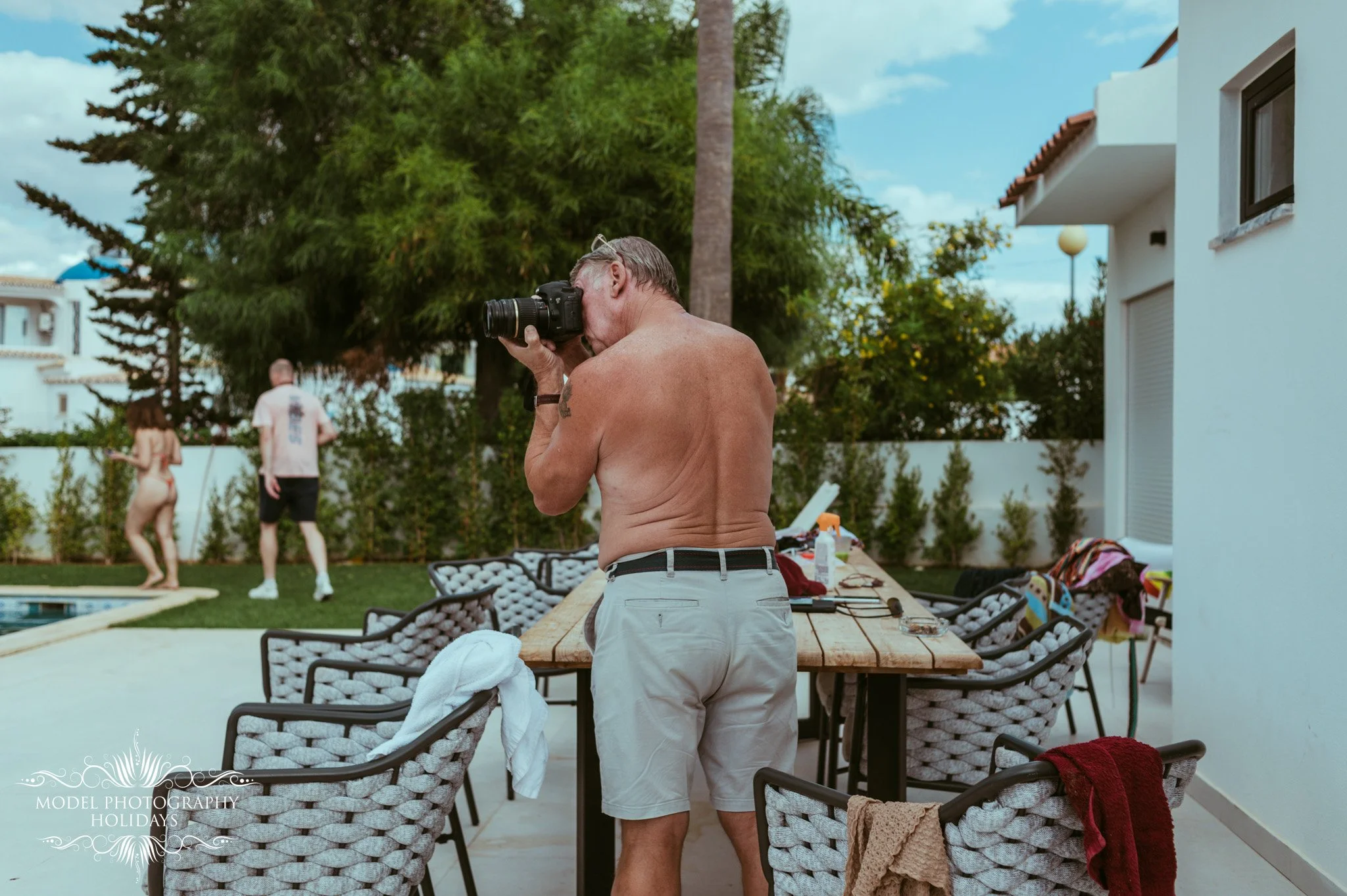 A shirtless man with gray hair taking a photograph with a digital camera outdoors on a patio area with tables and chairs, with two women in swimsuits and a white house with orange roof tiles in the background.