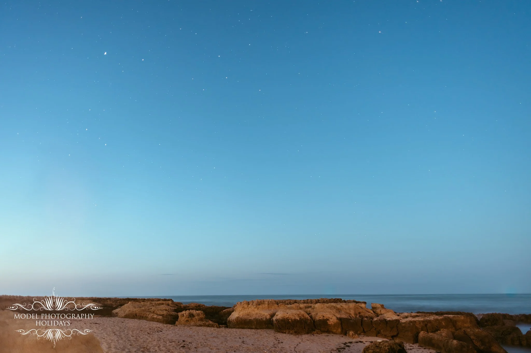Coastal landscape with rocky shoreline, calm ocean, clear blue sky with stars.