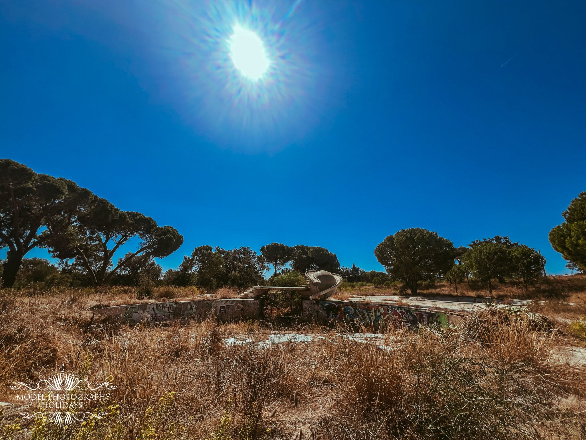 A bright sun in a clear blue sky above a field with brown dry grass, trees, and a graffiti-covered concrete structure.