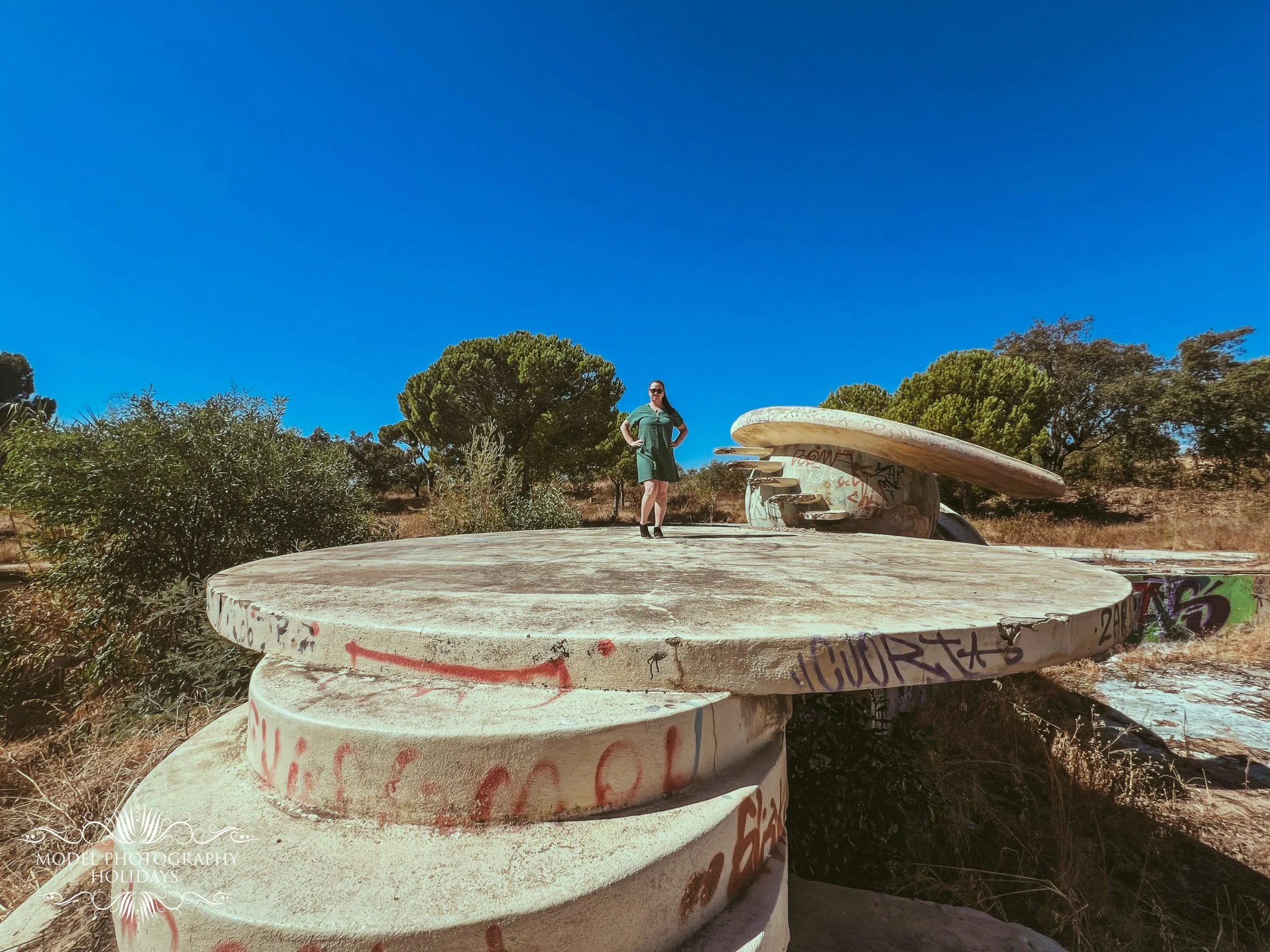 A woman with long dark hair and sunglasses standing on large, graffiti-covered concrete structures in a dry, tree-filled landscape under a bright blue sky.
