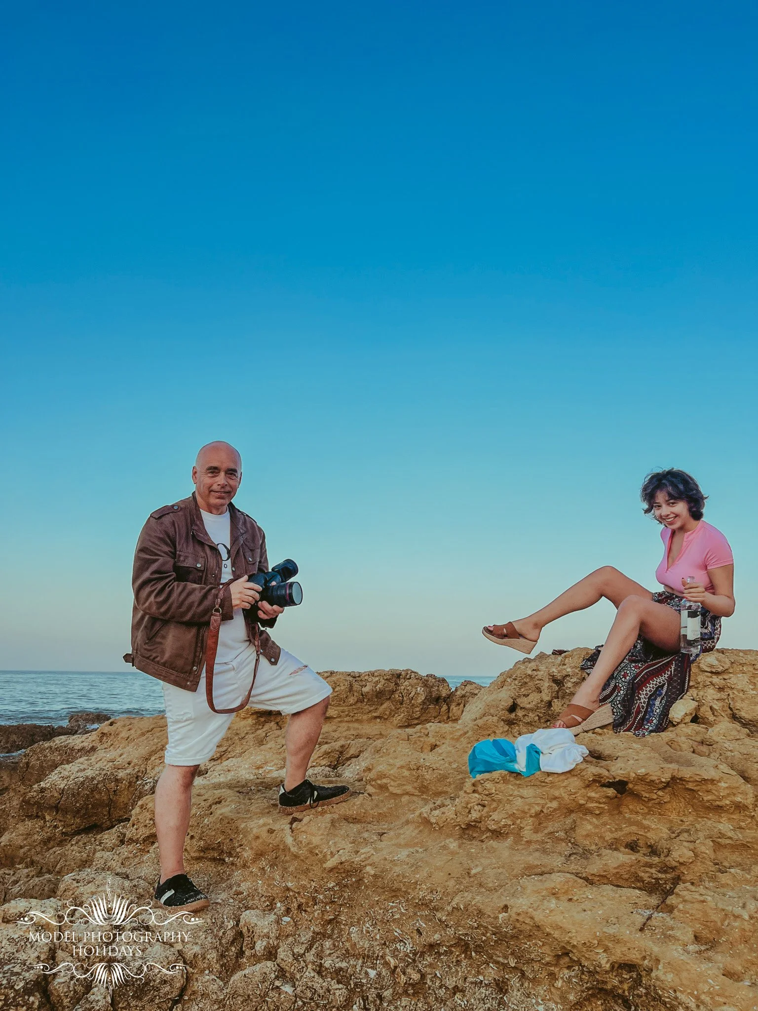 A man and a woman on a rocky seashore during sunset. The man is standing on the rocks, holding a camera, and the woman is sitting on the rocks, smiling and holding a bottle. The ocean is in the background under a clear sky.