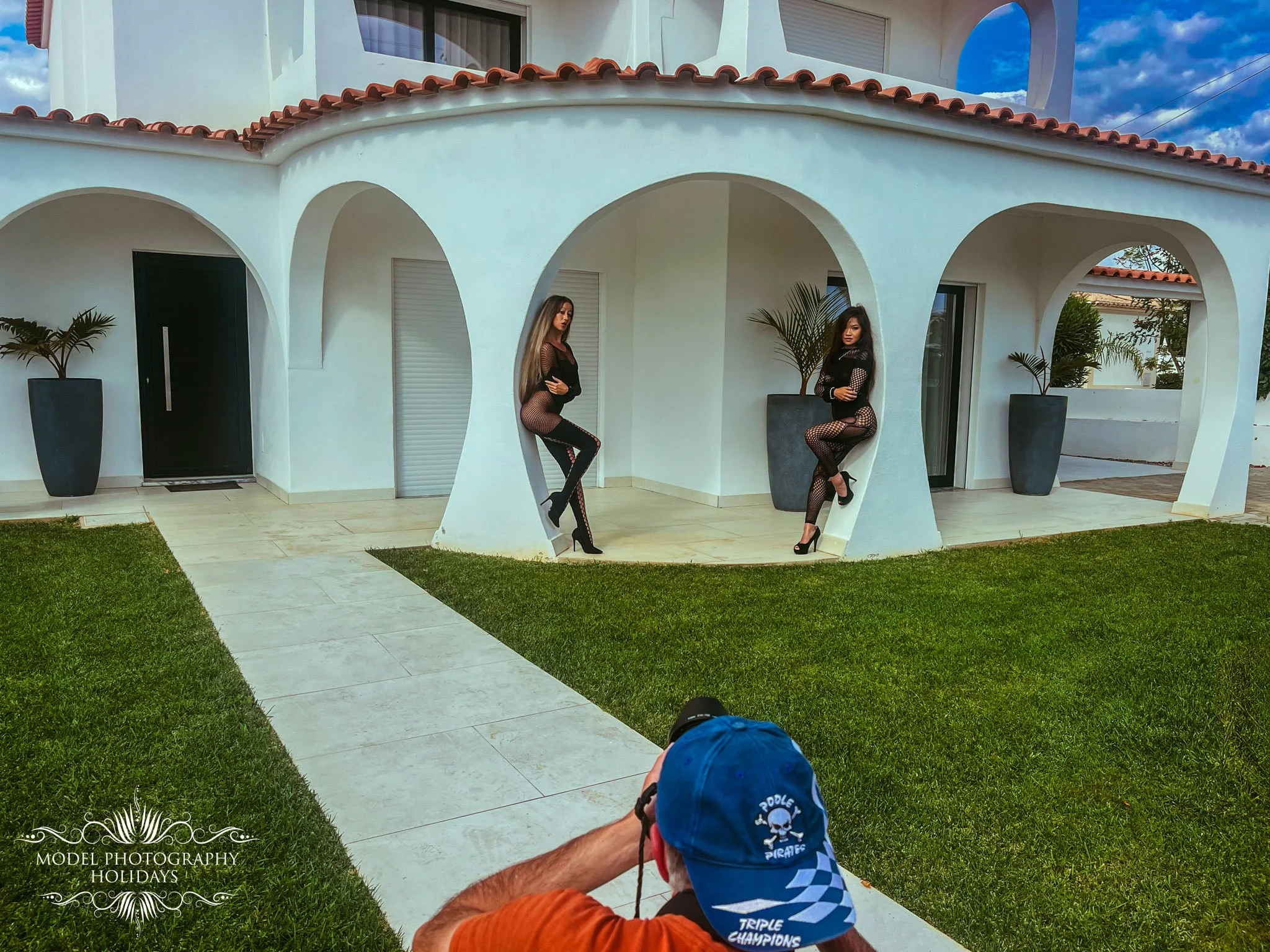 Two women in black fishnet outfits standing in arched opening of a white building with red tile roof, flanked by potted plants, with photographer capturing their photo.