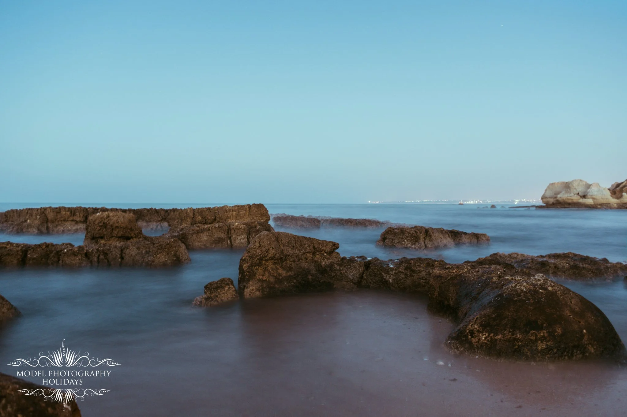 Calm ocean water with rocks in the foreground, a large rock formation in the distance on the right, and a clear blue sky above.
