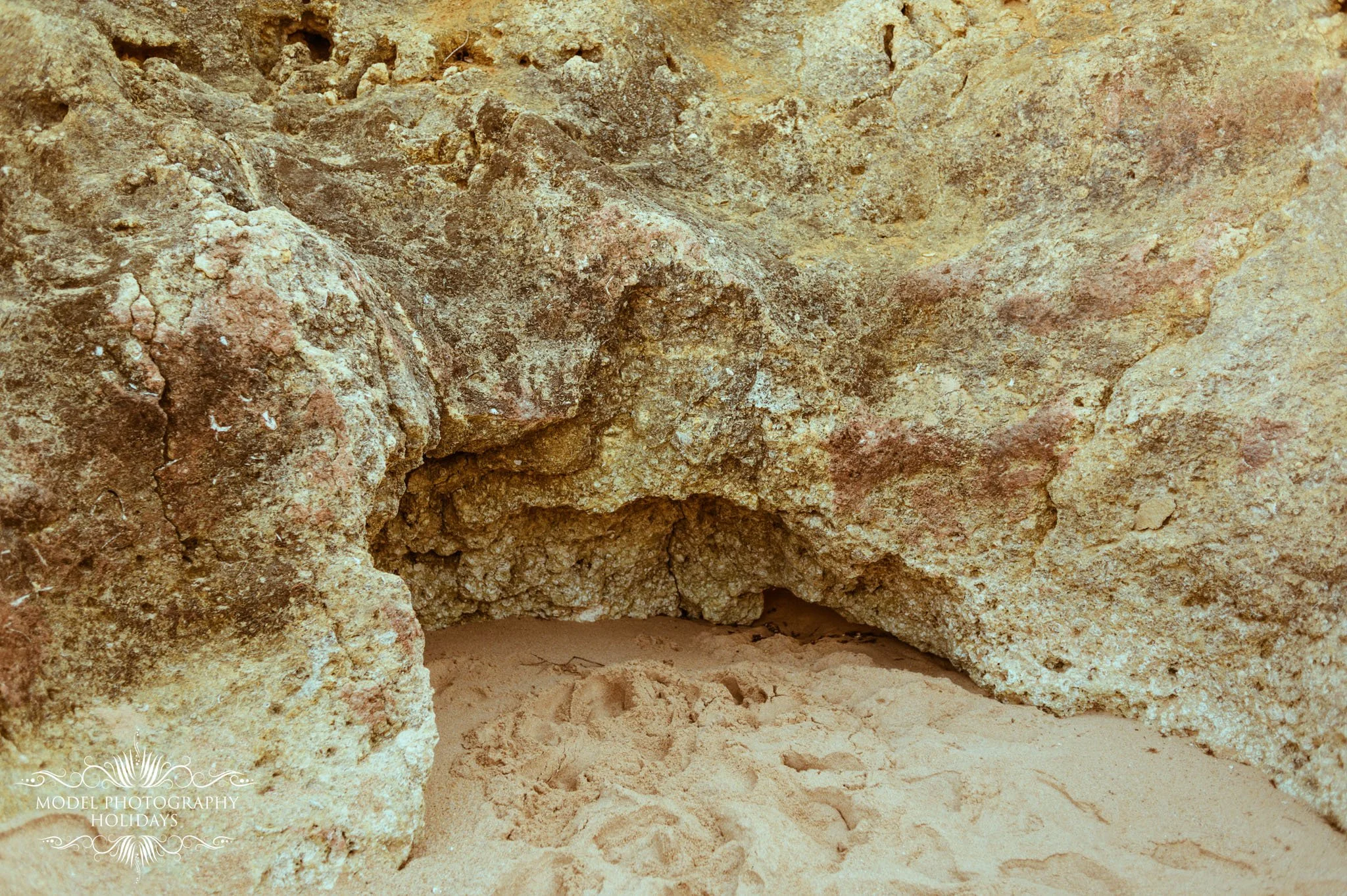 A close-up view of a cave entrance with rugged, textured, sandy-colored rock walls and a small, dark opening leading into darkness.