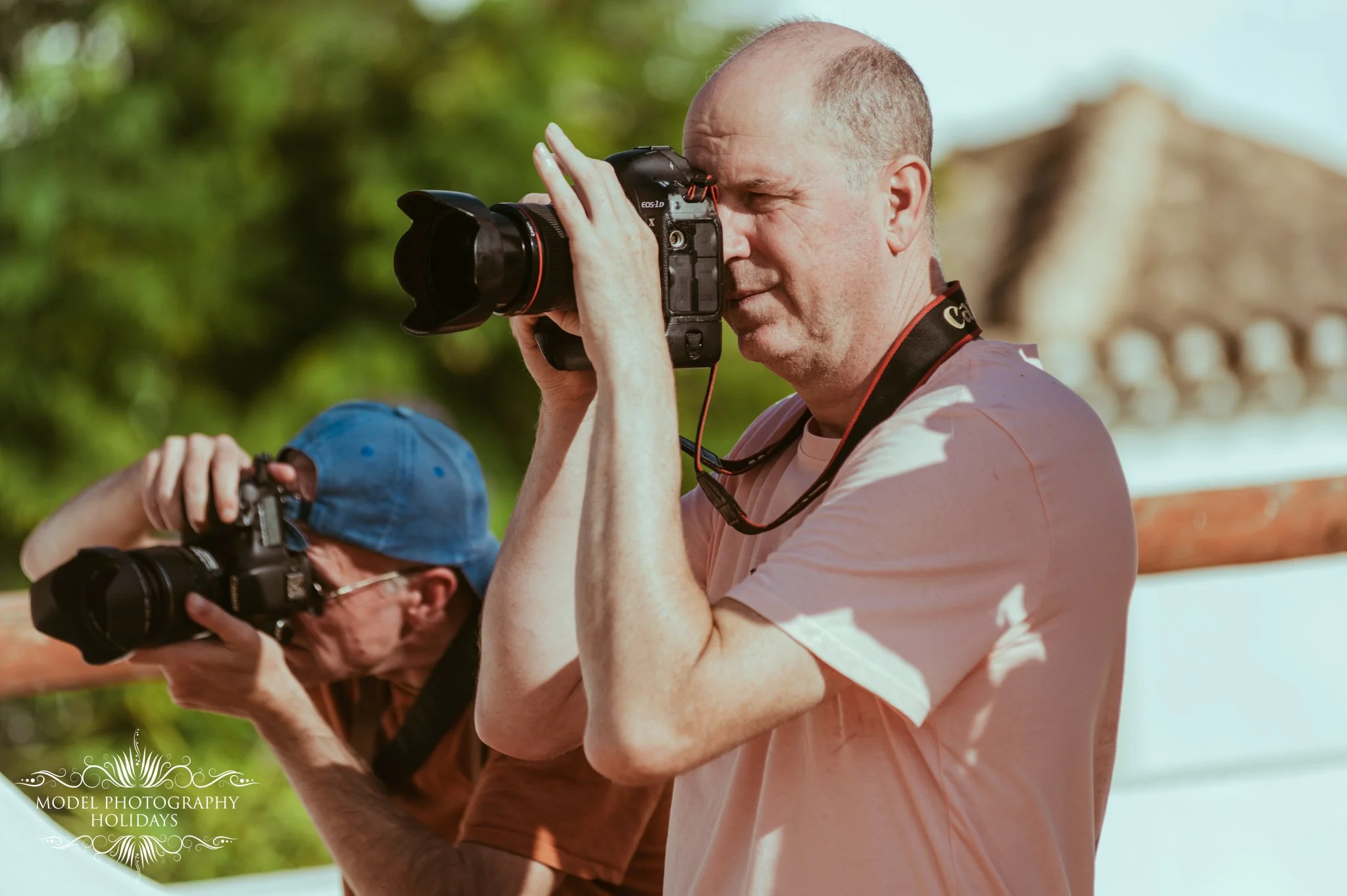 Two men are taking photographs outdoors. The man in the foreground is looking through the viewfinder of his camera, which has a large lens. The man in the background is also holding a camera and is focused on photographing something. Both are standin