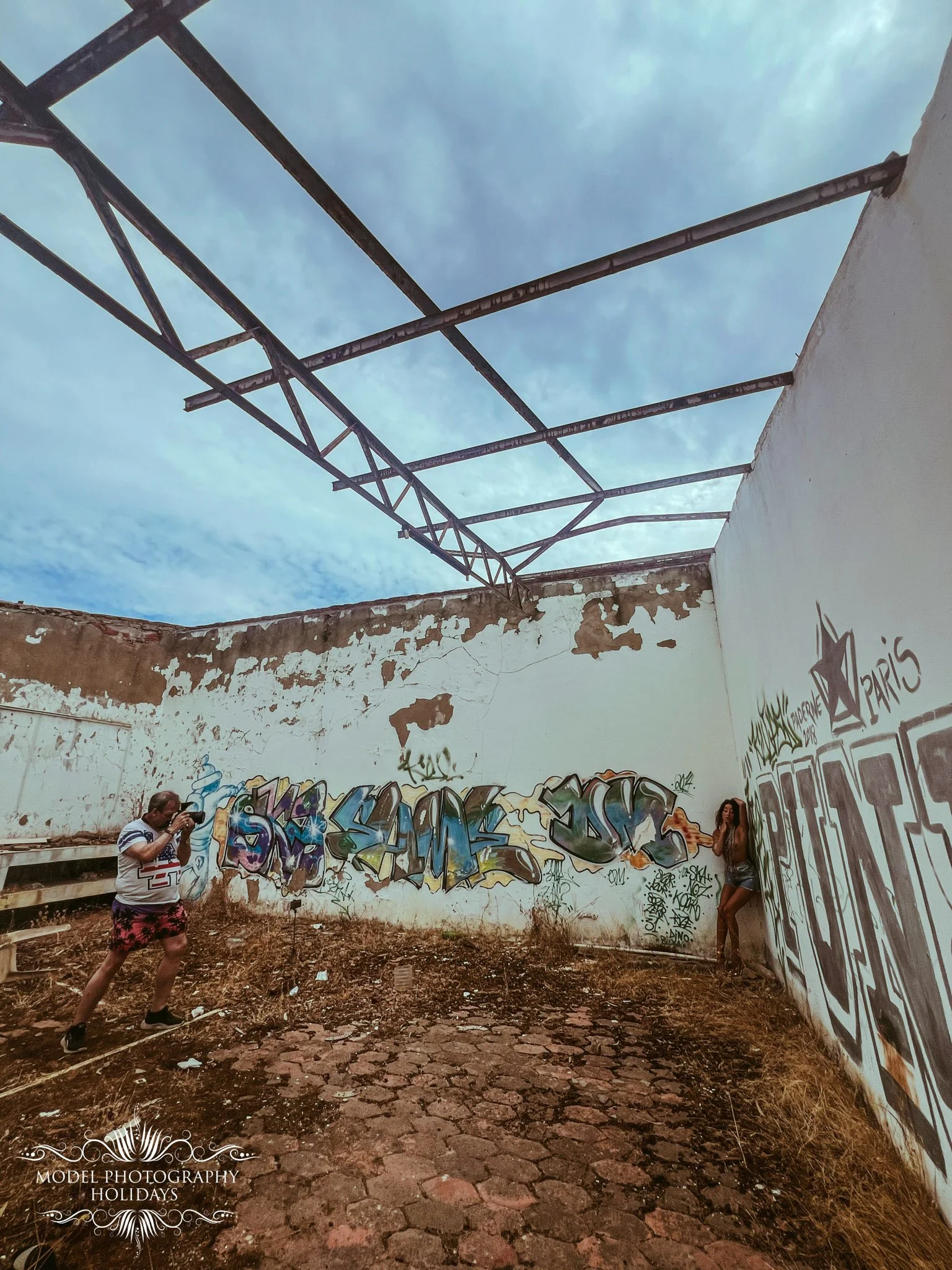 Two people in an abandoned, graffiti-covered building with a partially collapsed roof and dirt floor. One person is taking a photo and the other is leaning against the wall. The building has graffiti art on the walls, and the sky can be seen through 