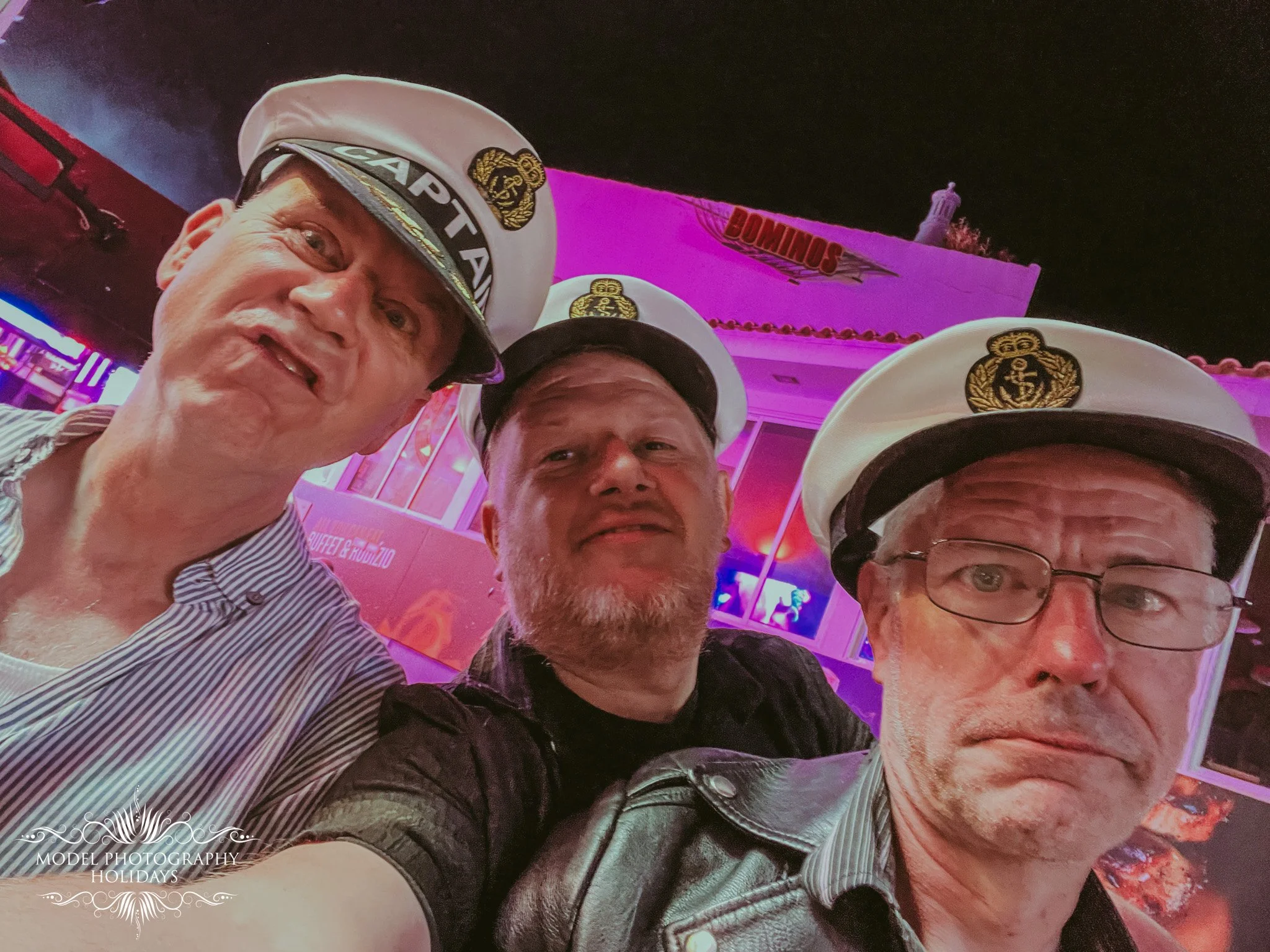 A group of four men, three wearing white sailor hats, taking a selfie in front of a building with pink and purple neon lighting at night.