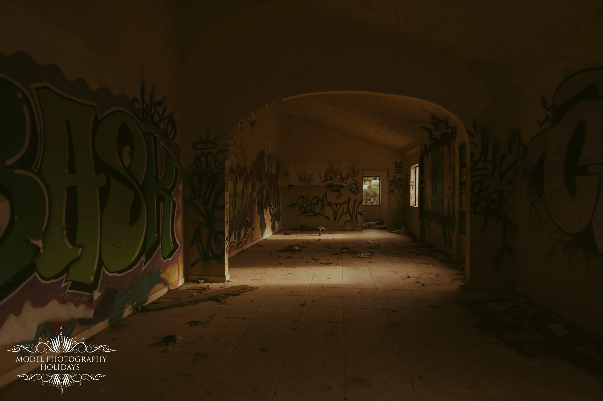 An abandoned building corridor with graffiti on the walls, broken tiles on the floor, and sunlight coming through open windows at the end.