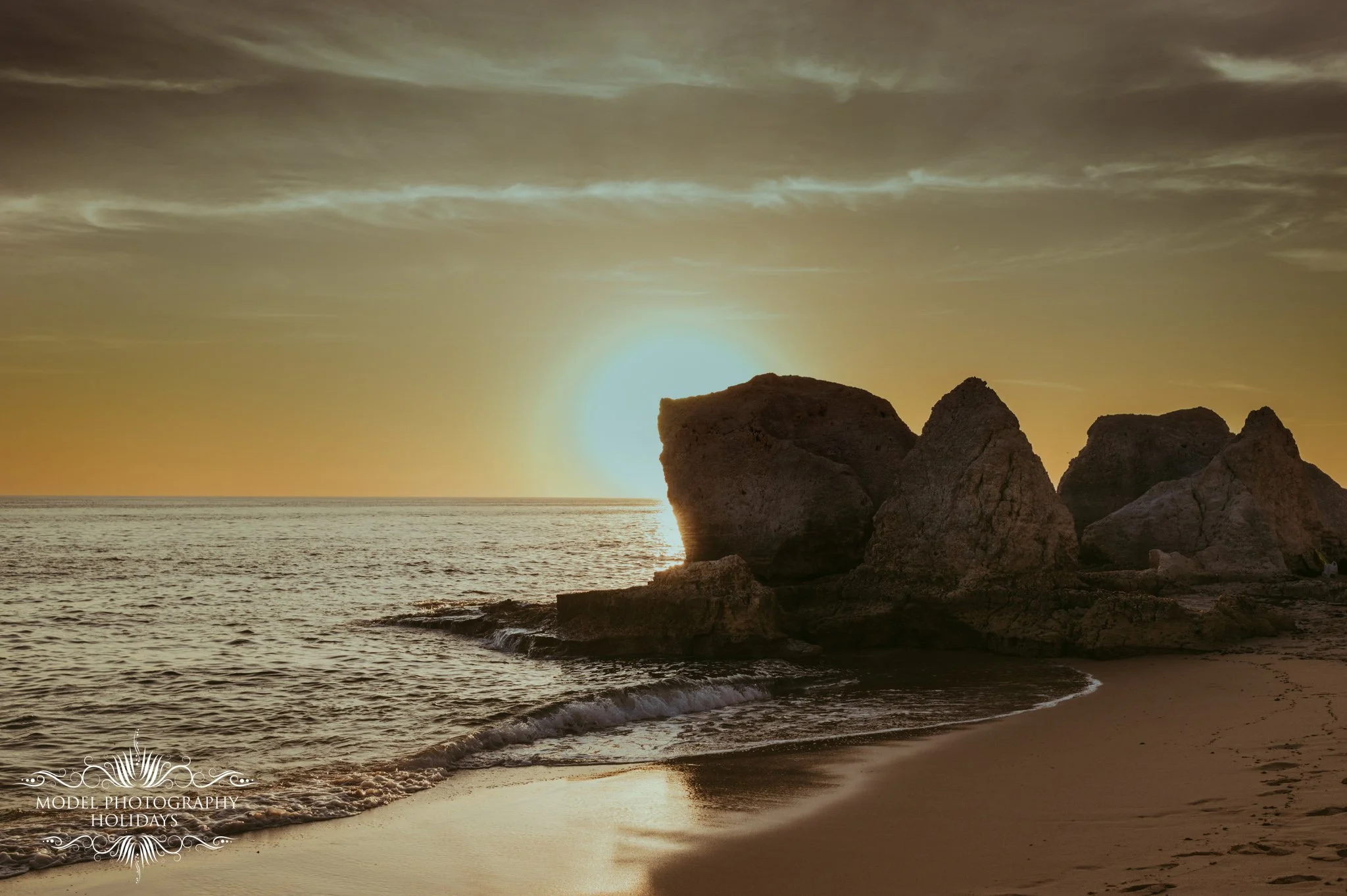 A sunset over the ocean with large rock formations on the shoreline, partly cloudy sky, and gentle waves on the beach.
