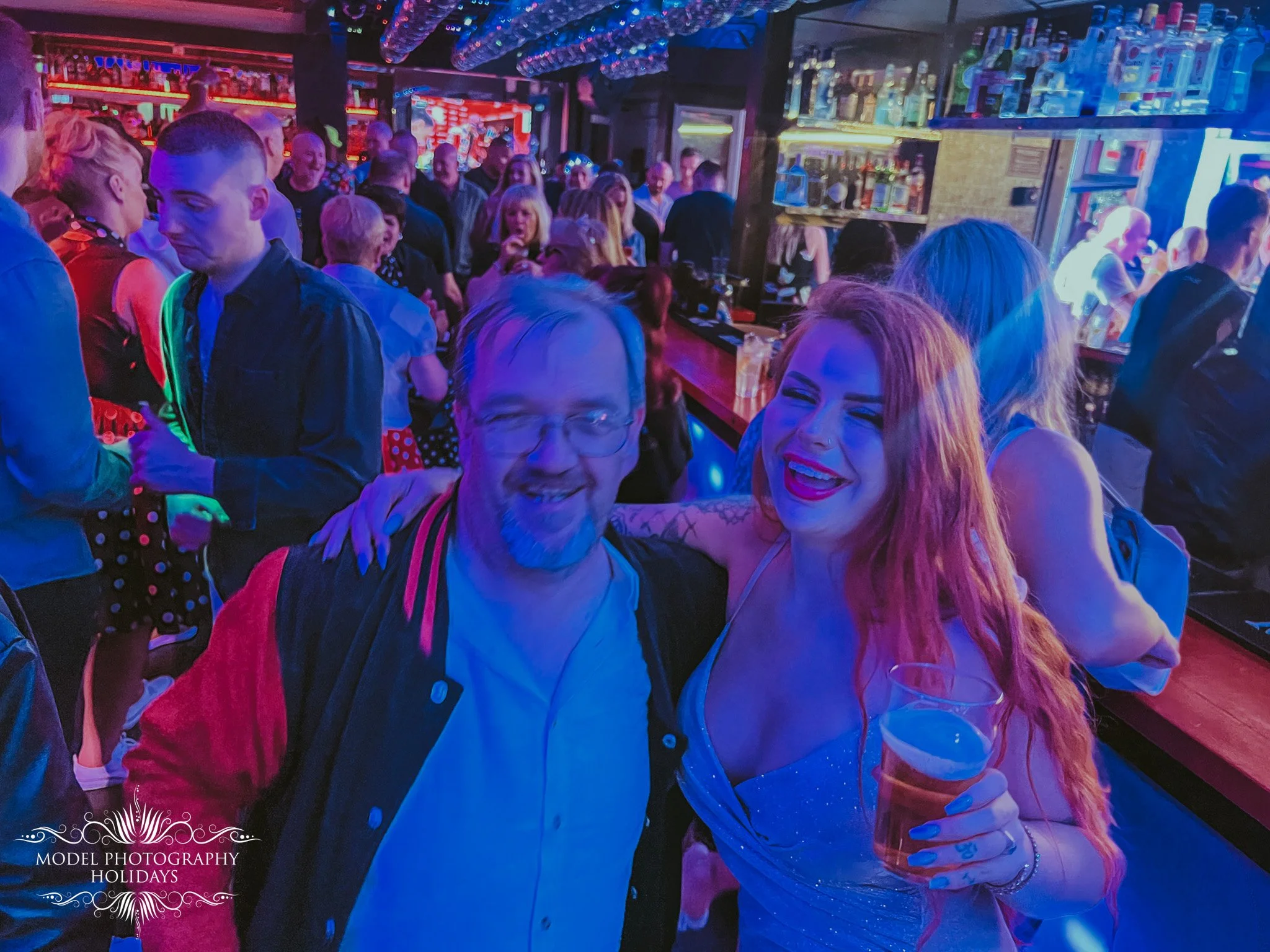 A crowded bar filled with people socializing and enjoying drinks. The room is dimly lit with colorful neon lighting, and shelves behind the bar are stocked with bottles of alcohol.