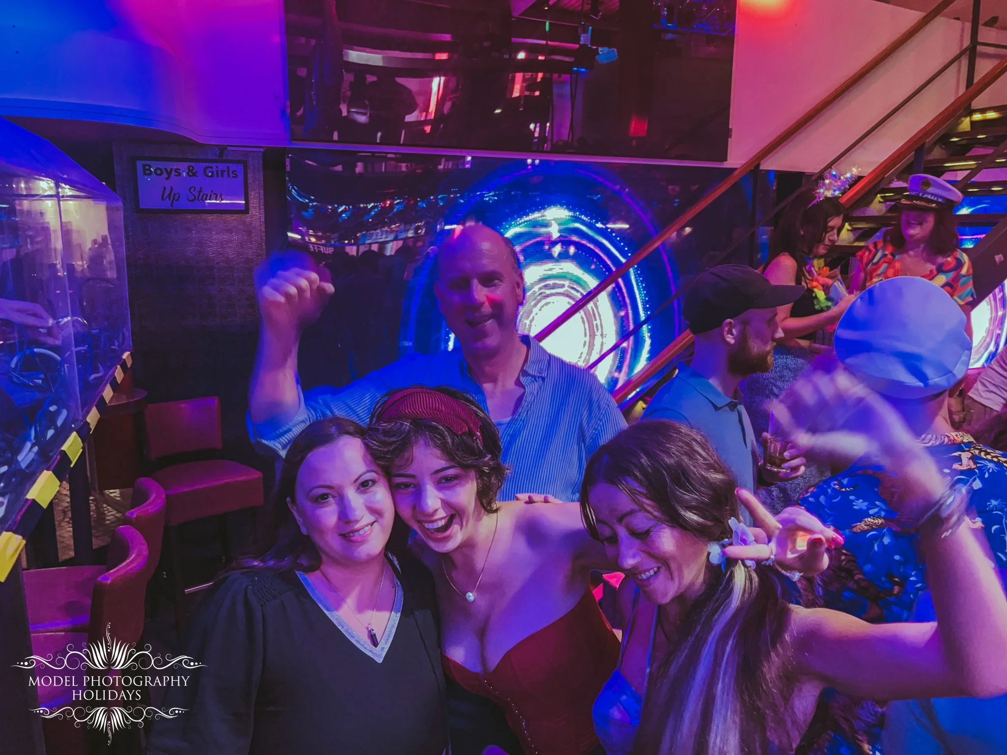 People taking a group photo at a lively indoor event with vibrant, colorful lighting, and a digital tunnel background, some wearing costumes and accessories like hats and balloons.
